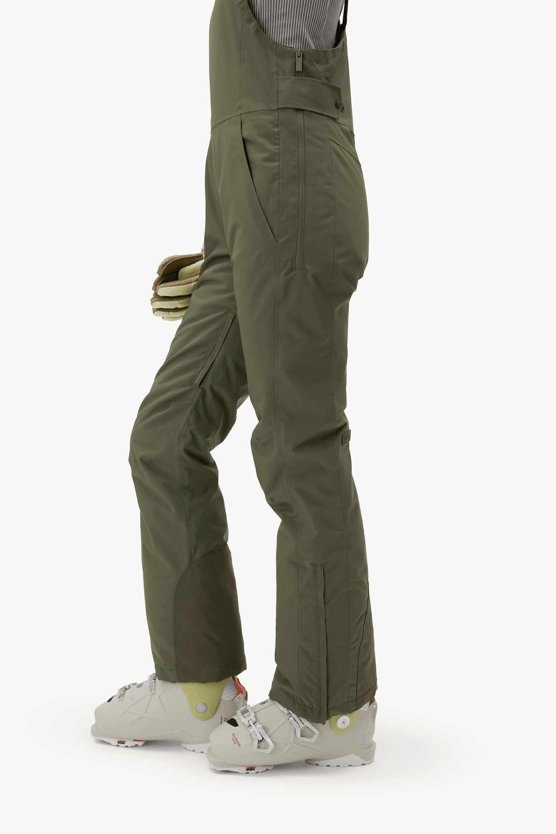 Side view of a person wearing olive green ski bib pants with white ski boots, shown against a plain light background.