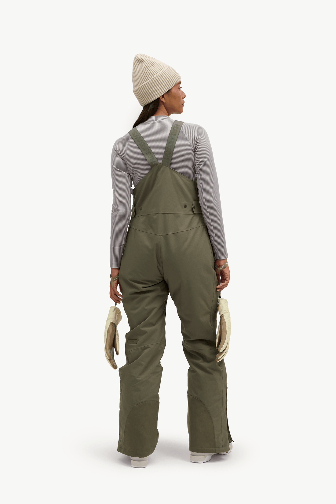 A model from behind wearing olive green snow bib overalls over a light gray long-sleeve shirt, a cream knit beanie, and holding a pair of cream-colored gloves against a solid off-white background.