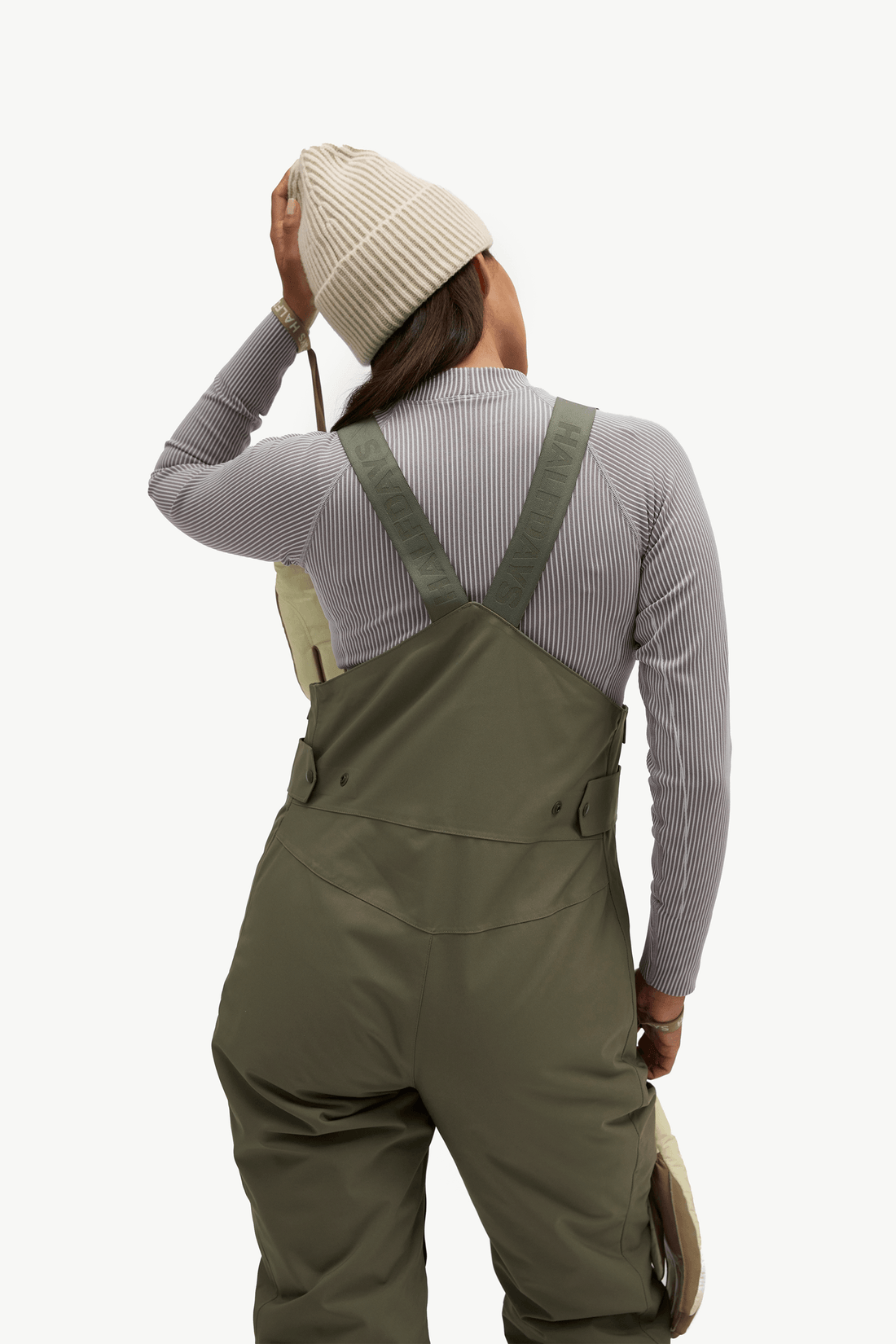 A person shown from the back wearing olive green bib overalls over a light gray ribbed long-sleeve top, with a cream knit beanie and a light tan hat visible, against a plain light background.