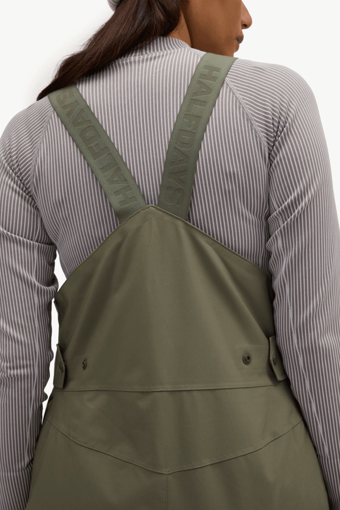 A person shown from the back wearing olive green overall-style bib overalls over a gray and white vertically striped long-sleeve shirt. The straps have visible text reading 'HALFTAYE'.