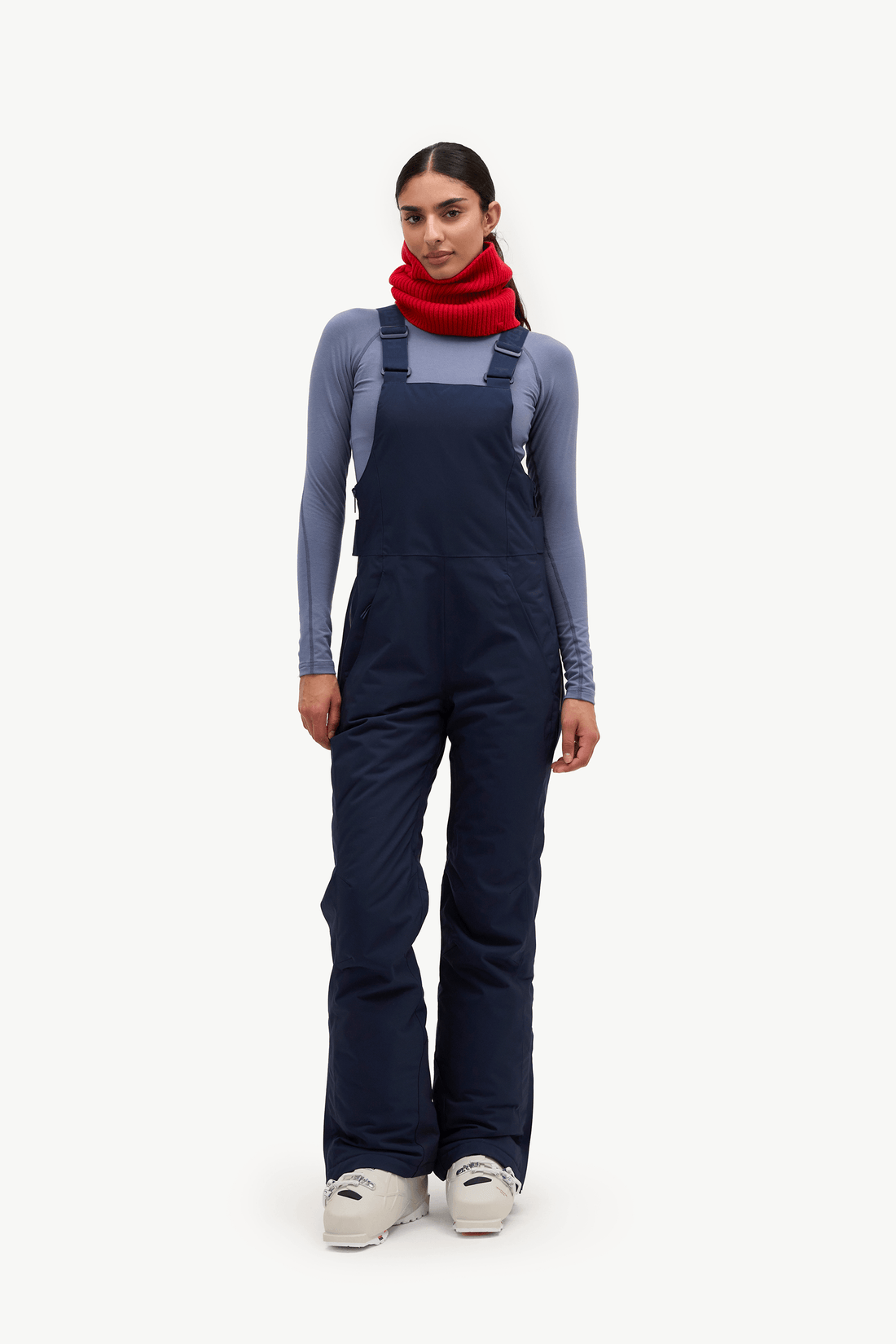 A model stands against a plain white studio background wearing dark navy insulated bib overalls over a slate blue long-sleeve top, a bright red scarf, and white sneakers.