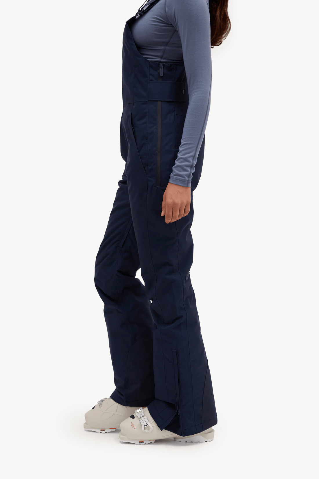 A woman modeling navy blue snow bib overalls over a slate blue long-sleeve shirt, accessorized with a bright red knit neck warmer, against a solid off-white background.