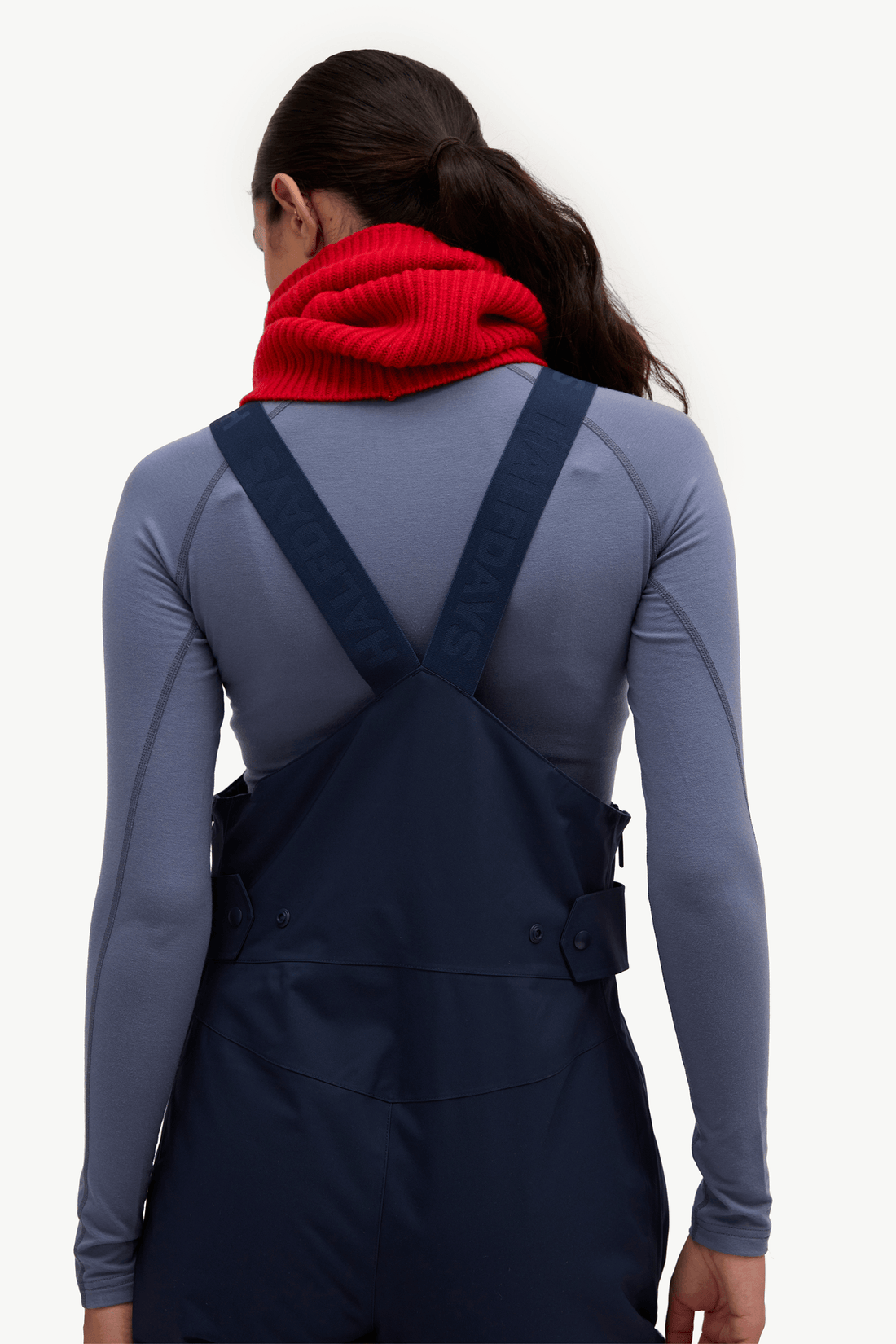 A woman modeling navy blue snow bib overalls over a slate blue long-sleeve shirt, accessorized with a bright red knit neck warmer, against a solid off-white background.