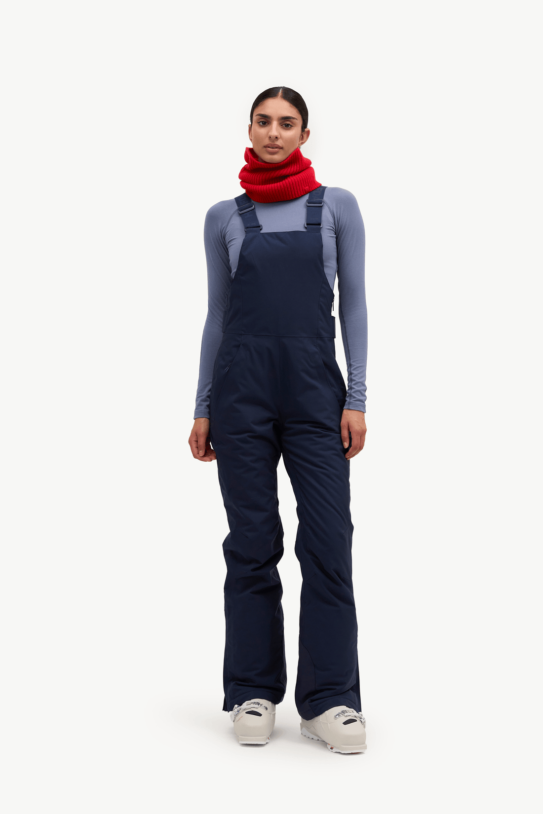 A model stands against a light gray studio background wearing dark navy insulated bib overalls over a slate blue-gray long-sleeve top, a bright red knit neck warmer, and light gray sneakers.