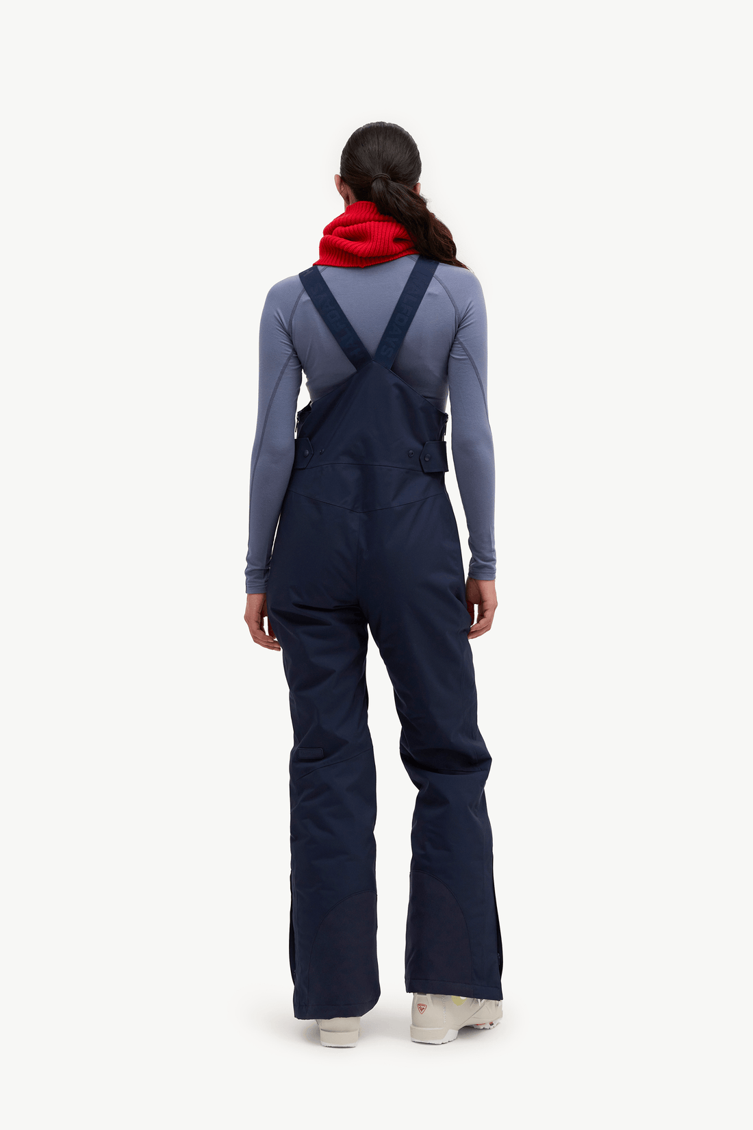 A model viewed from the back wearing navy blue snow bib overalls over a slate blue-gray long-sleeve top and a bright red neck warmer, standing against a solid off-white background.