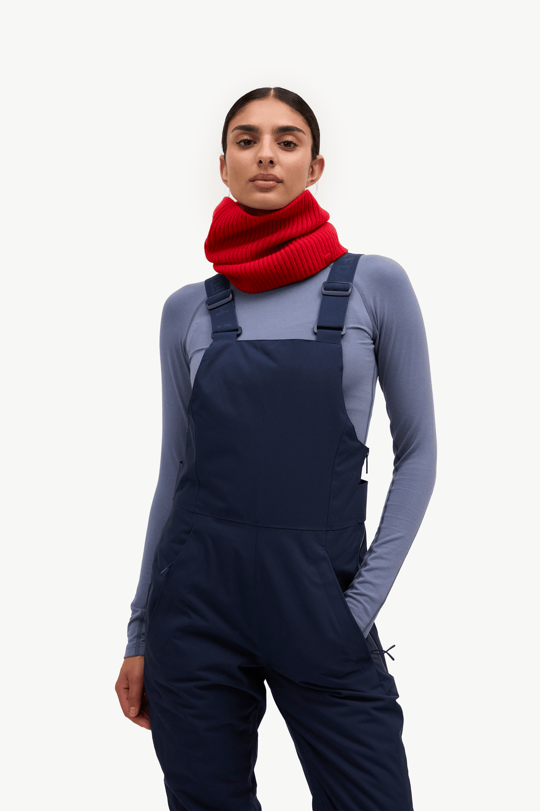 A woman modeling navy blue snow bib overalls over a slate blue long-sleeve shirt, accessorized with a bright red knit neck warmer, against a solid off-white background.
