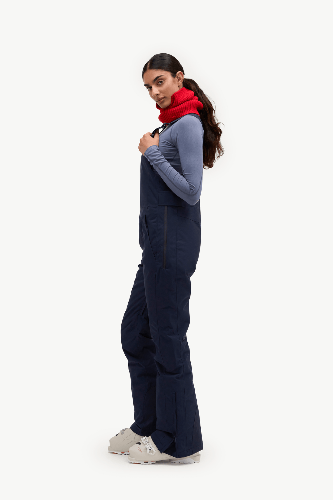 A woman modeling navy blue snow bib overalls over a slate blue long-sleeve shirt, accessorized with a bright red knit neck warmer, against a solid off-white background.