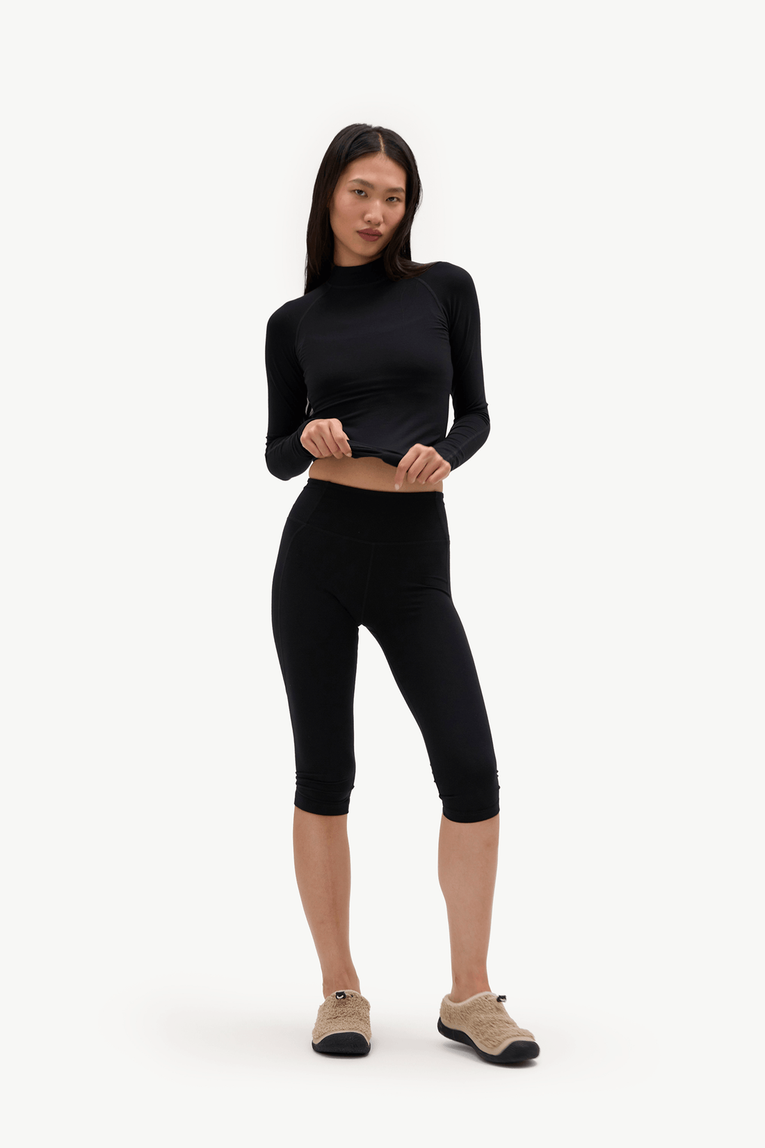 A woman modeling a black long-sleeve cropped mock neck top and black capri leggings, paired with beige slip-on shoes, against a plain white studio background.