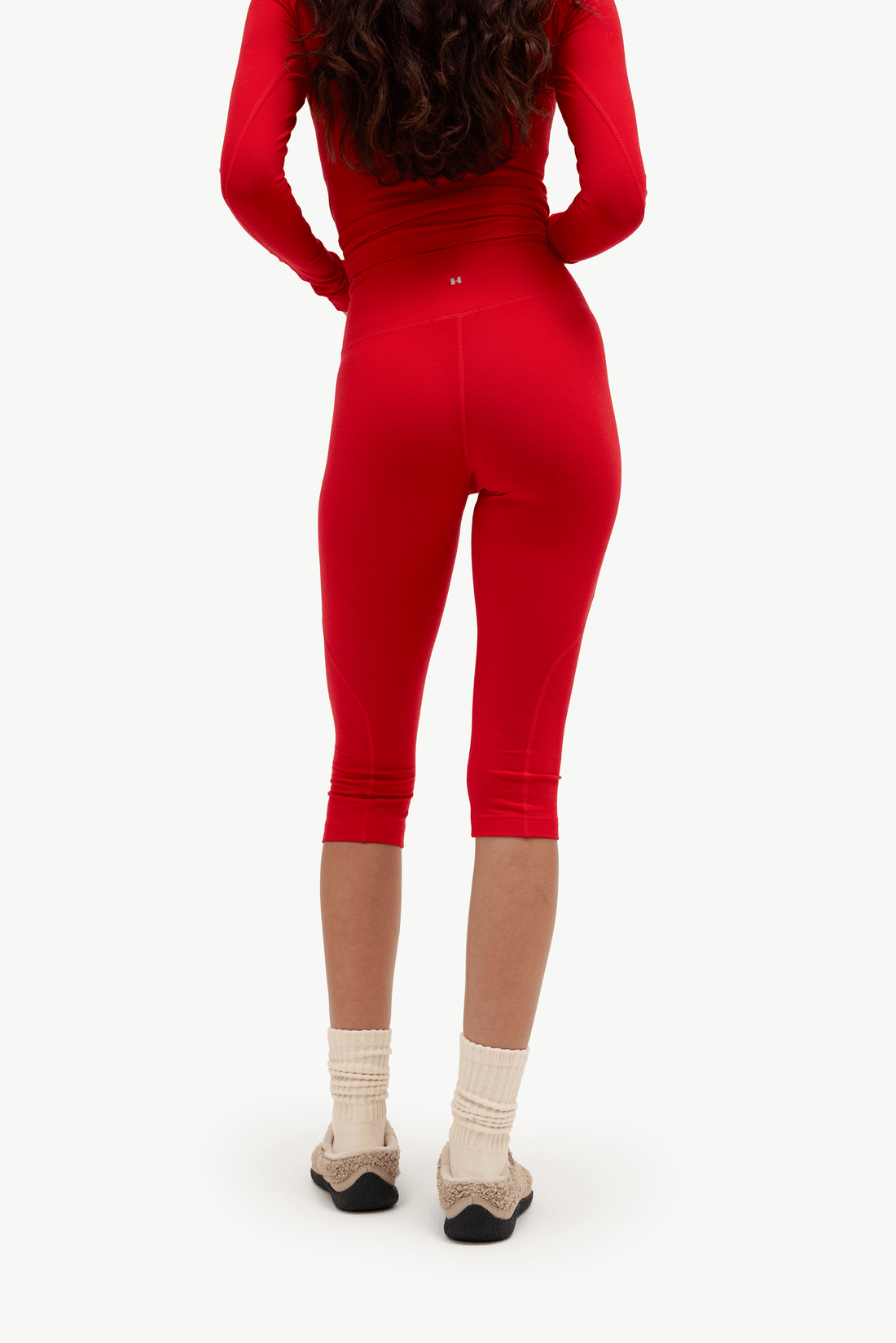 Rear view of a model wearing a bright red ribbed long-sleeve top and matching red capri leggings, paired with cream socks and beige flats, against a plain white background.