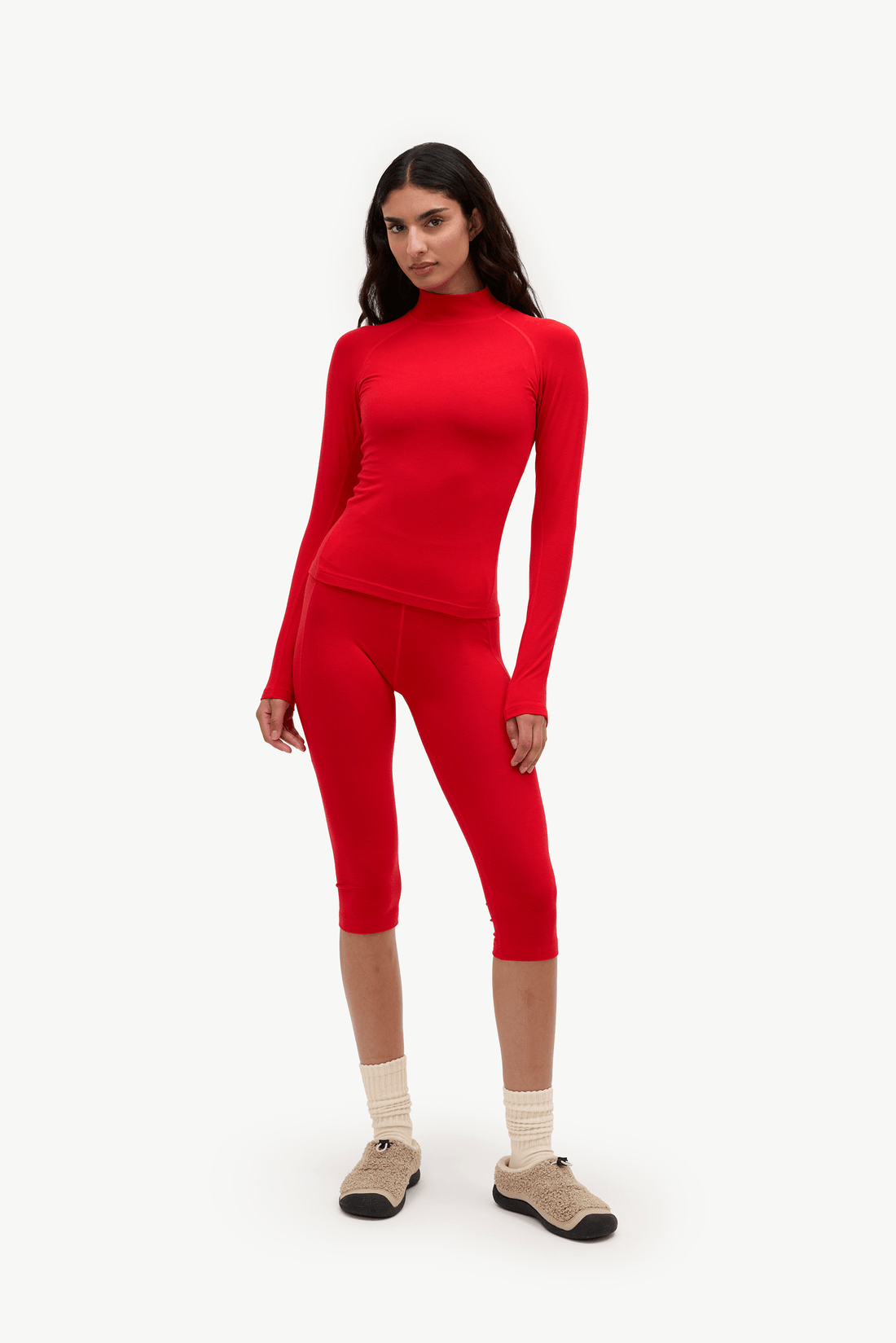 A woman modeling a matching bright red two-piece knit set consisting of a long-sleeve mock neck top and high-waisted capri leggings, styled with beige socks and brown slip-on shoes against a plain white background.