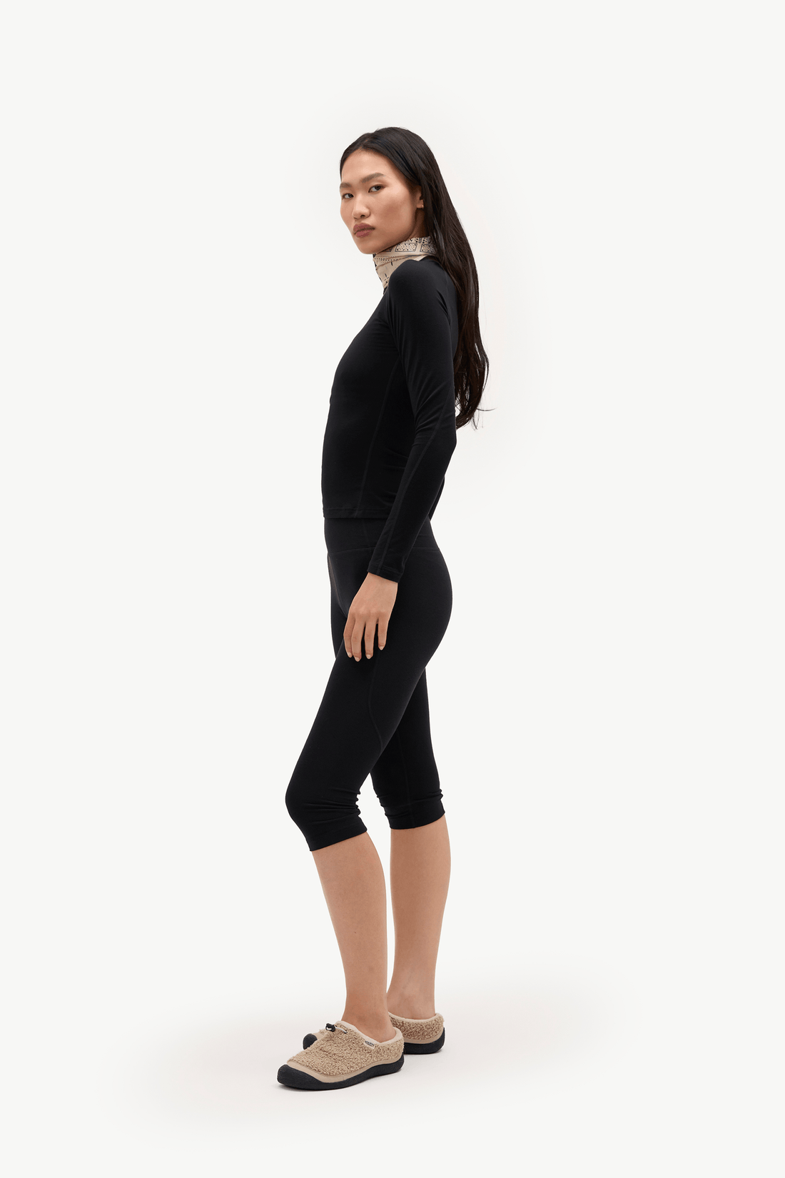 A woman modeling a black long-sleeve mock neck top and matching black capri leggings, paired with beige slip-on shoes, against a plain white studio background.
