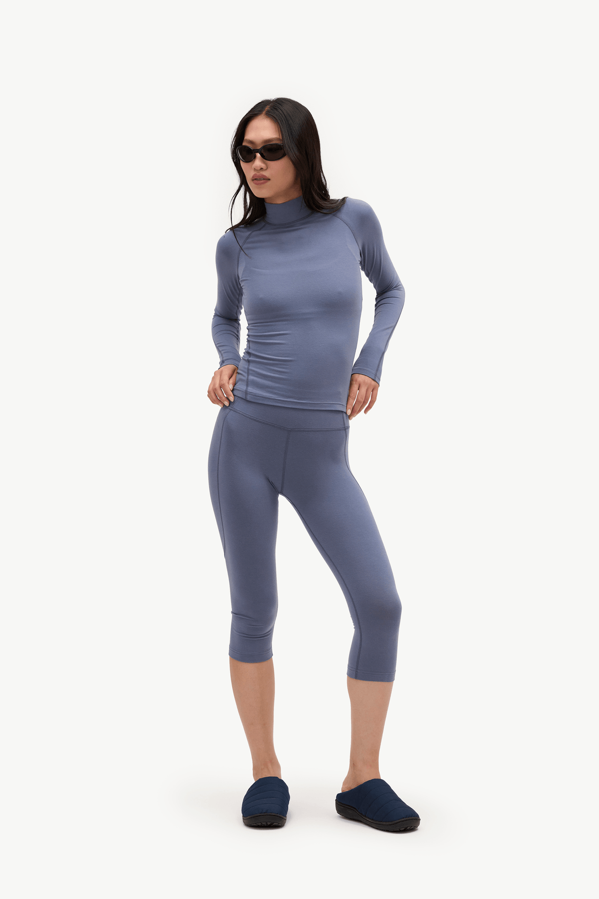 A woman modeling a matching two-piece activewear set consisting of a long-sleeve mock neck top and matching capri leggings, in a dusty slate blue color, paired with dark blue slip-on shoes against a plain white background.