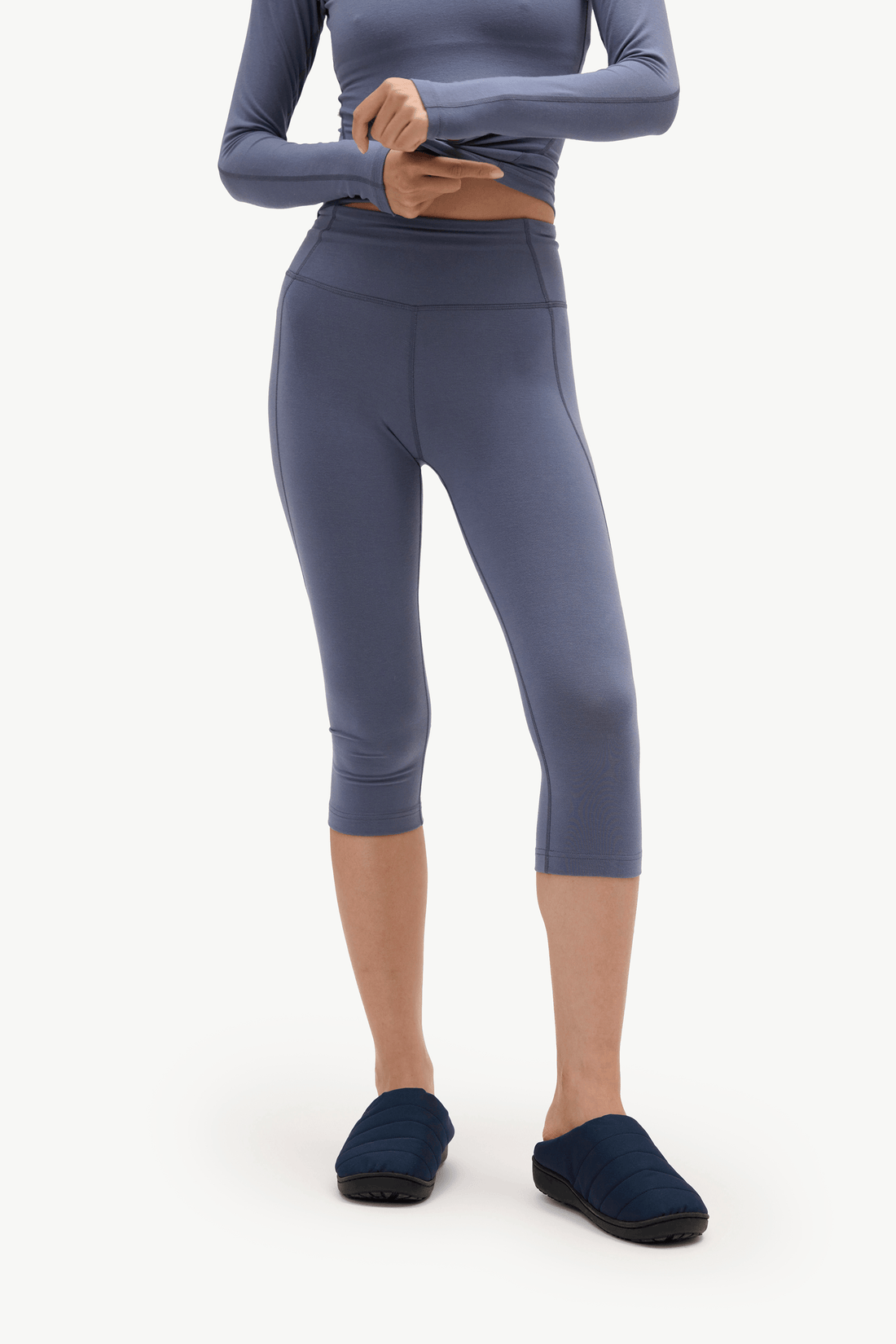 A model wearing slate blue-gray capri leggings, a matching long-sleeve crop top, and navy slip-on slippers, shown from the neck down against a plain white studio background.