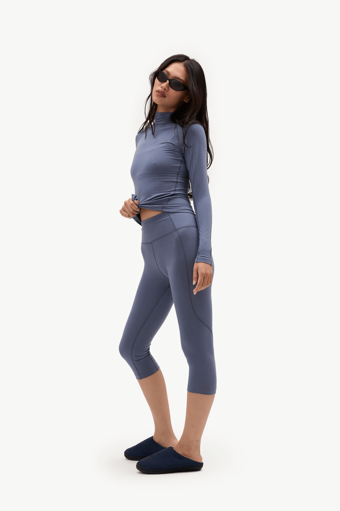 A woman modeling a matching two-piece activewear set consisting of a long-sleeve cropped mock-neck top and high-waisted capri leggings, in a dusty slate blue color. She is wearing dark sunglasses and dark slip-on shoes against a plain white studio background.