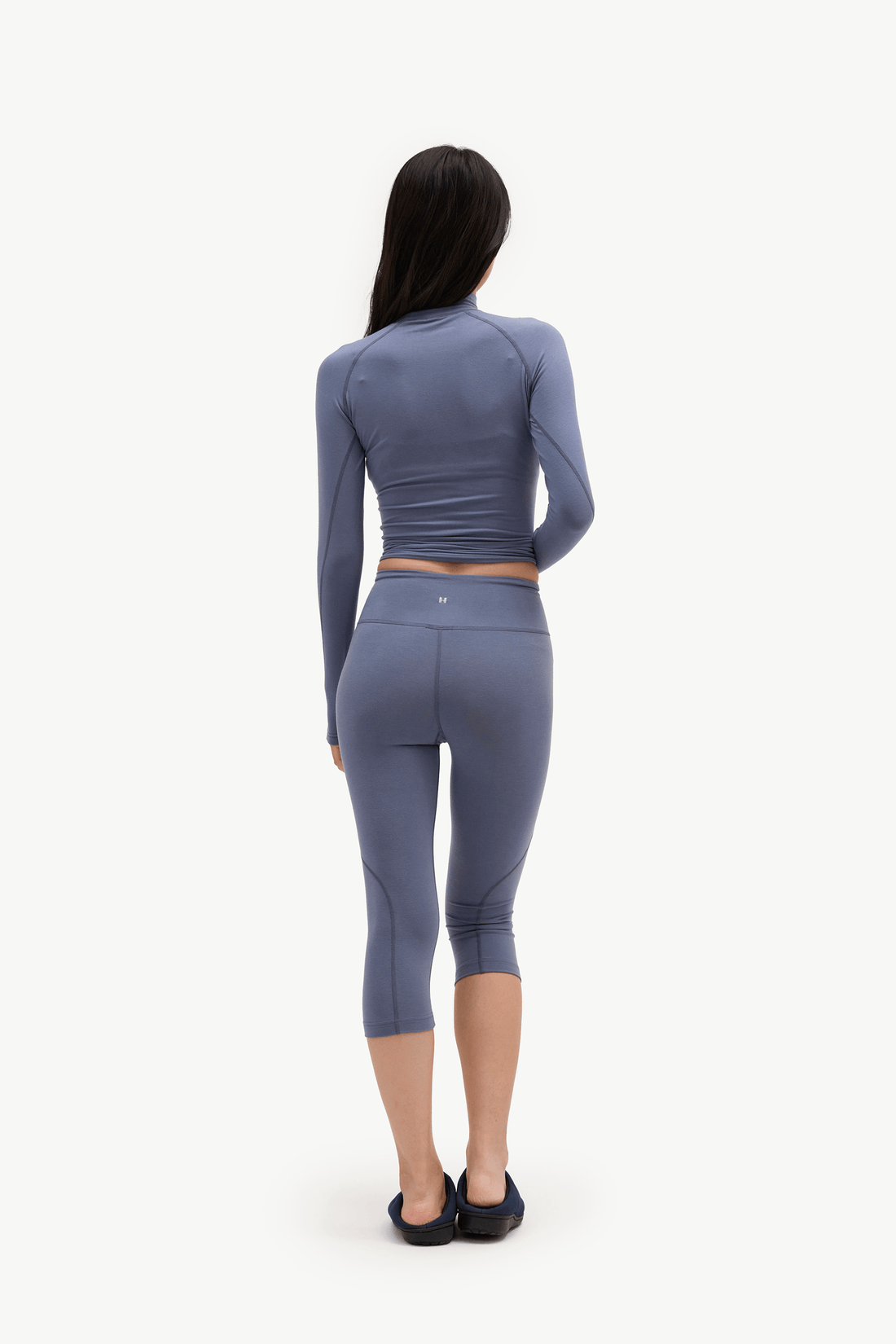 A woman viewed from the back wearing a matching slate blue-gray activewear set consisting of a long-sleeve crop top and capri leggings, paired with black flats, against a plain white studio background.