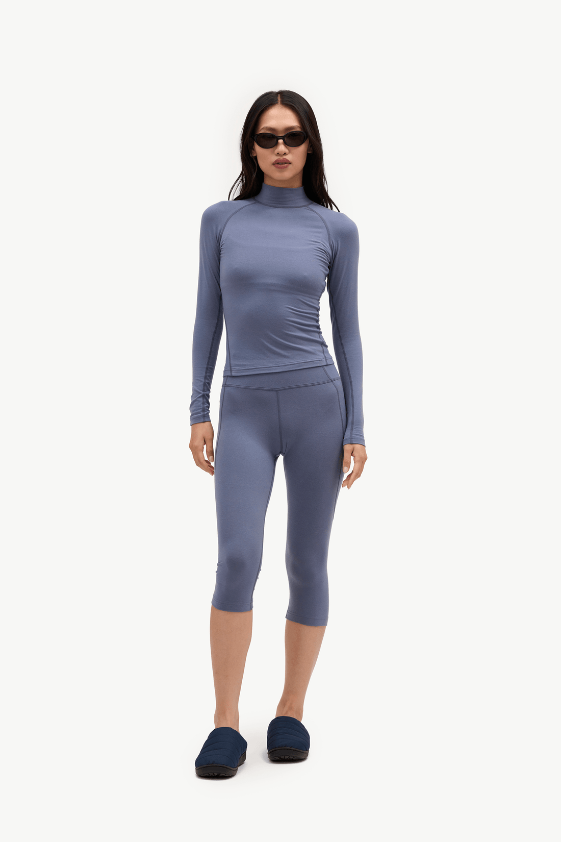 A woman modeling a matching two-piece activewear set consisting of a long-sleeve mock neck top and matching capri leggings in a dusty slate blue color, paired with dark blue slip-on shoes against a plain white background.