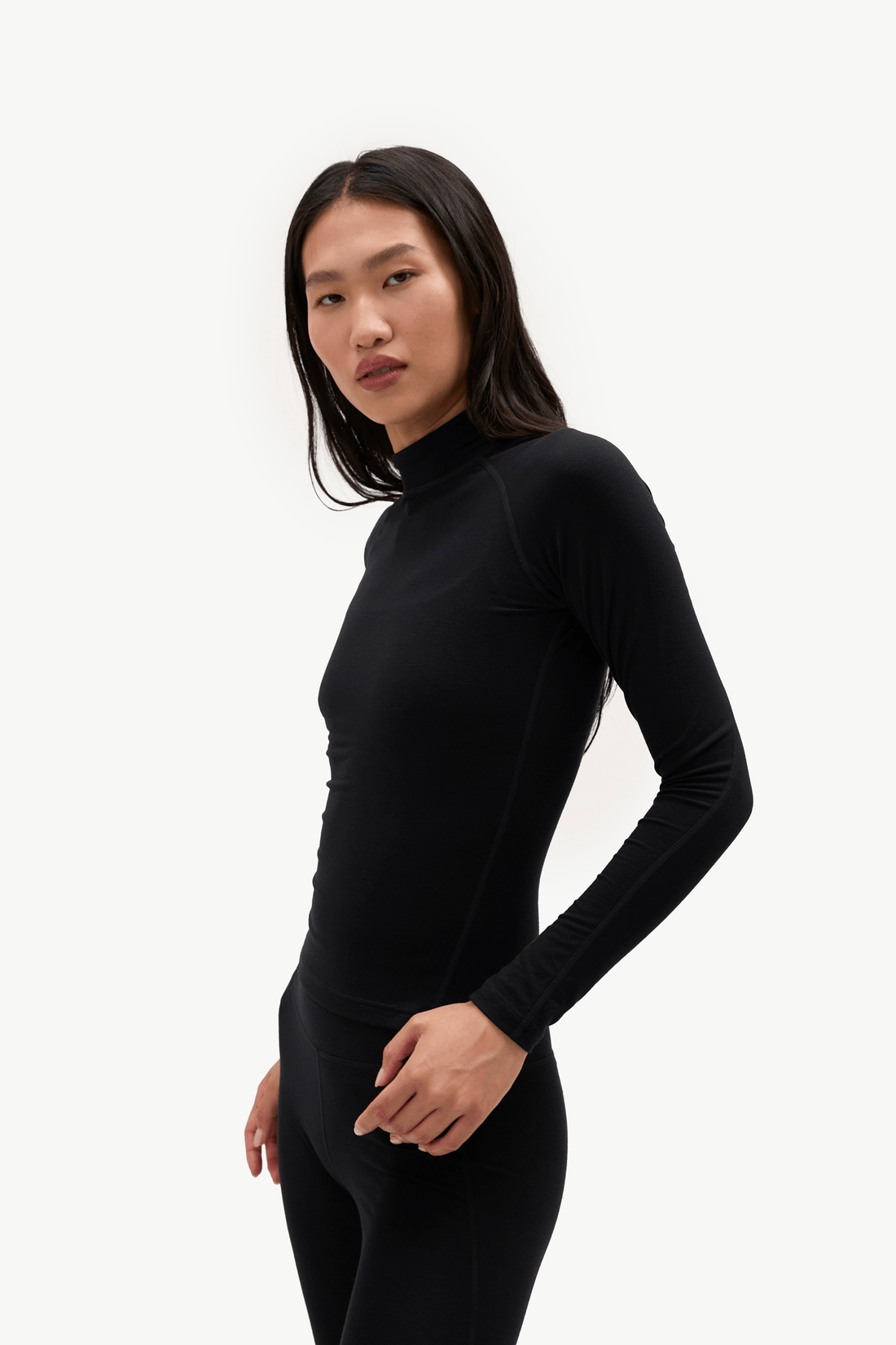 A woman modeling a black long-sleeve mock neck athletic top and matching black leggings against a plain white studio background.