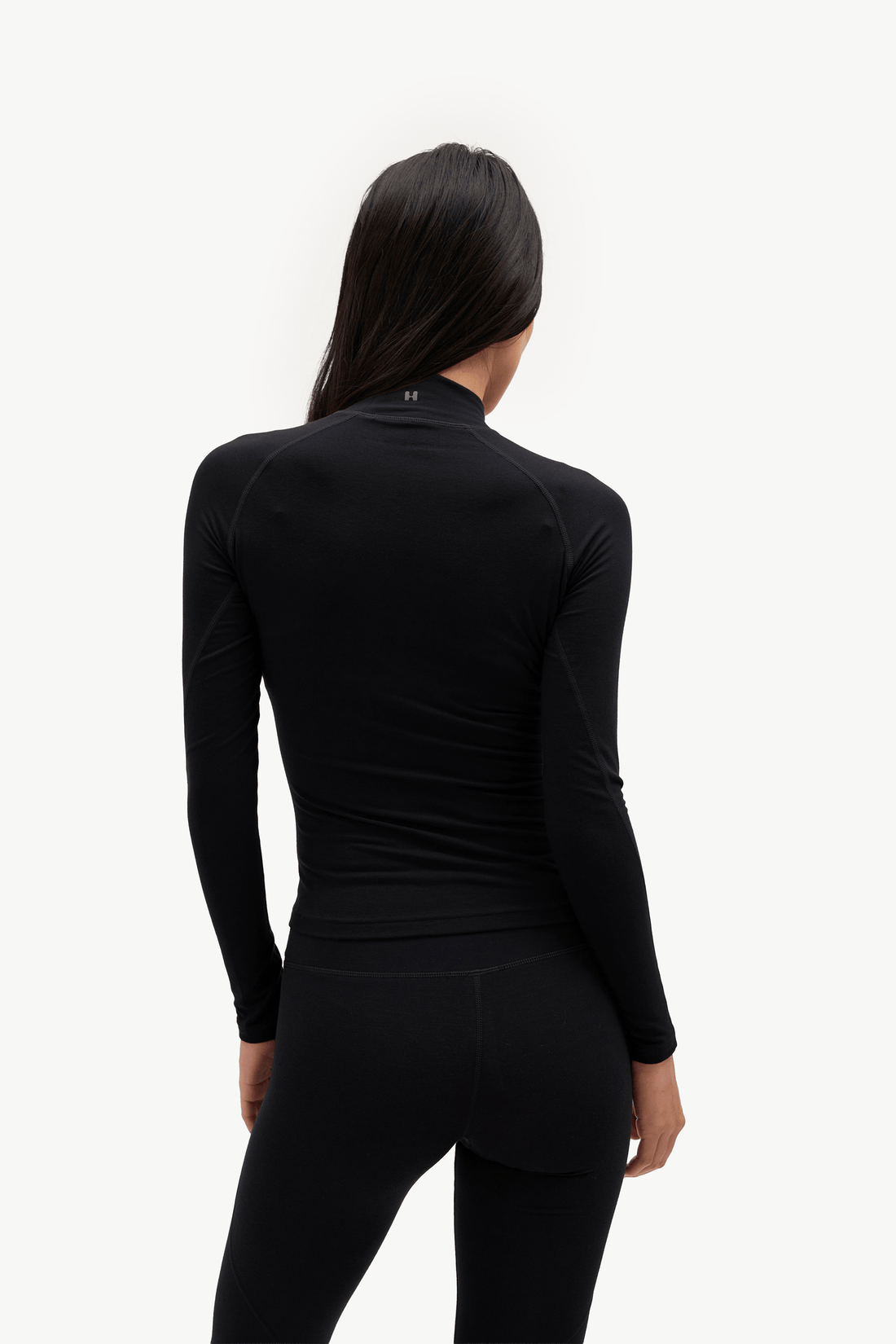 A woman viewed from the back wearing a fitted black long-sleeve athletic top and black leggings against a plain light background.