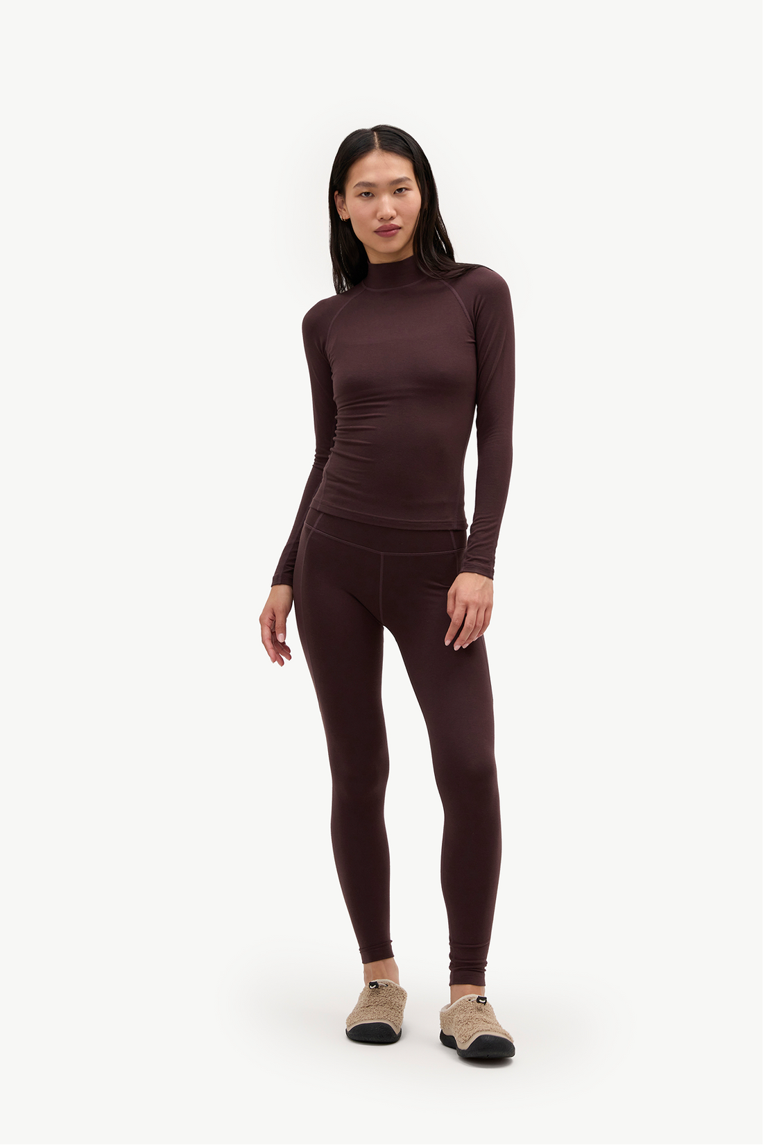 A woman modeling a matching dark brown long-sleeve mock neck top and high-waisted leggings set, paired with beige sneakers, against a plain white studio background.