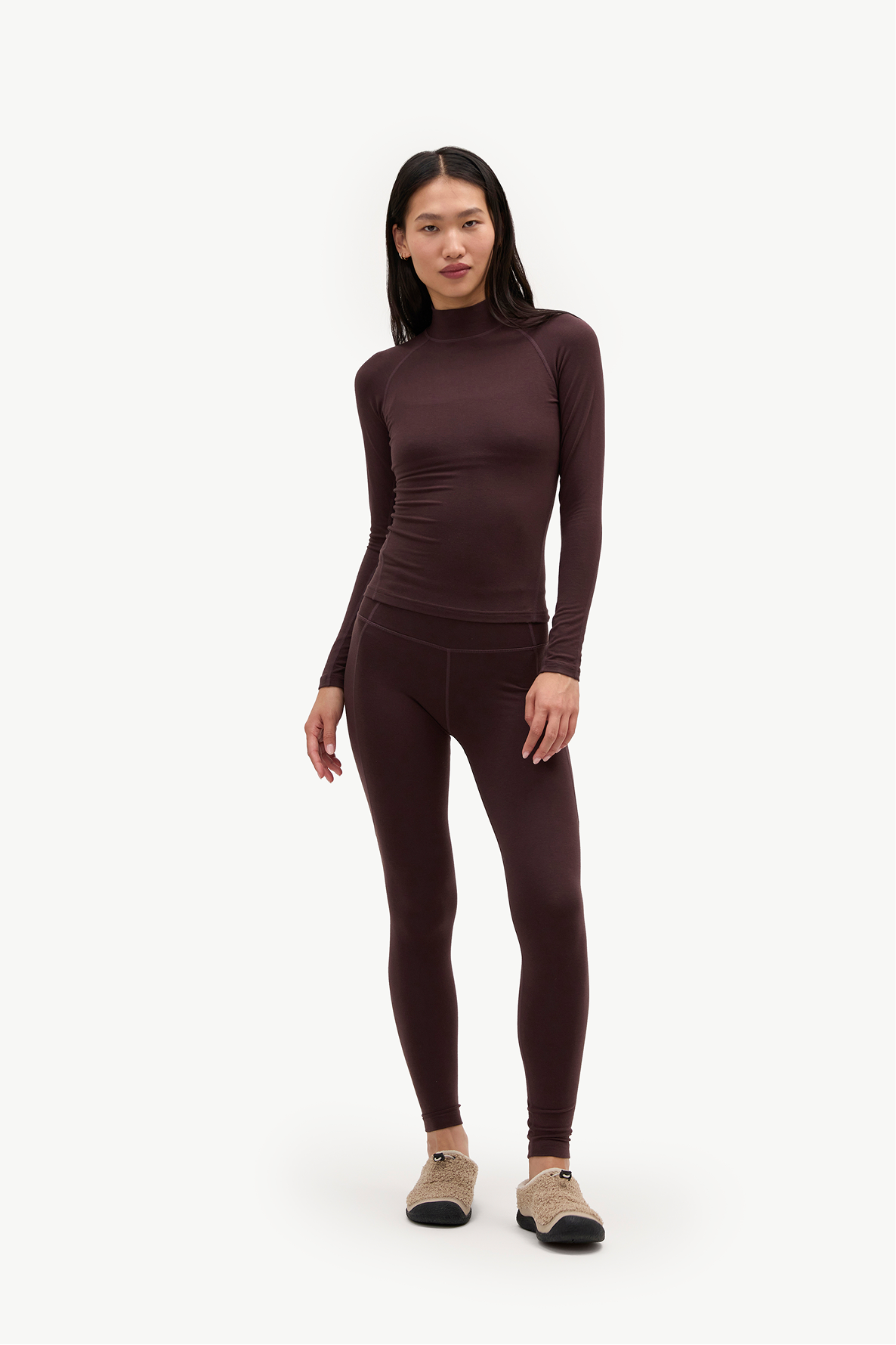 A woman modeling a matching dark brown long-sleeve mock neck top and high-waisted leggings set, paired with beige sneakers, against a plain white studio background.