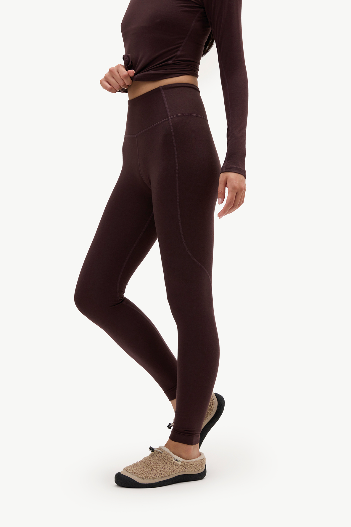 A model wearing dark brown high-waisted leggings and a matching long-sleeve crop top, paired with beige slip-on shoes, shown from the waist down against a light gray studio background.