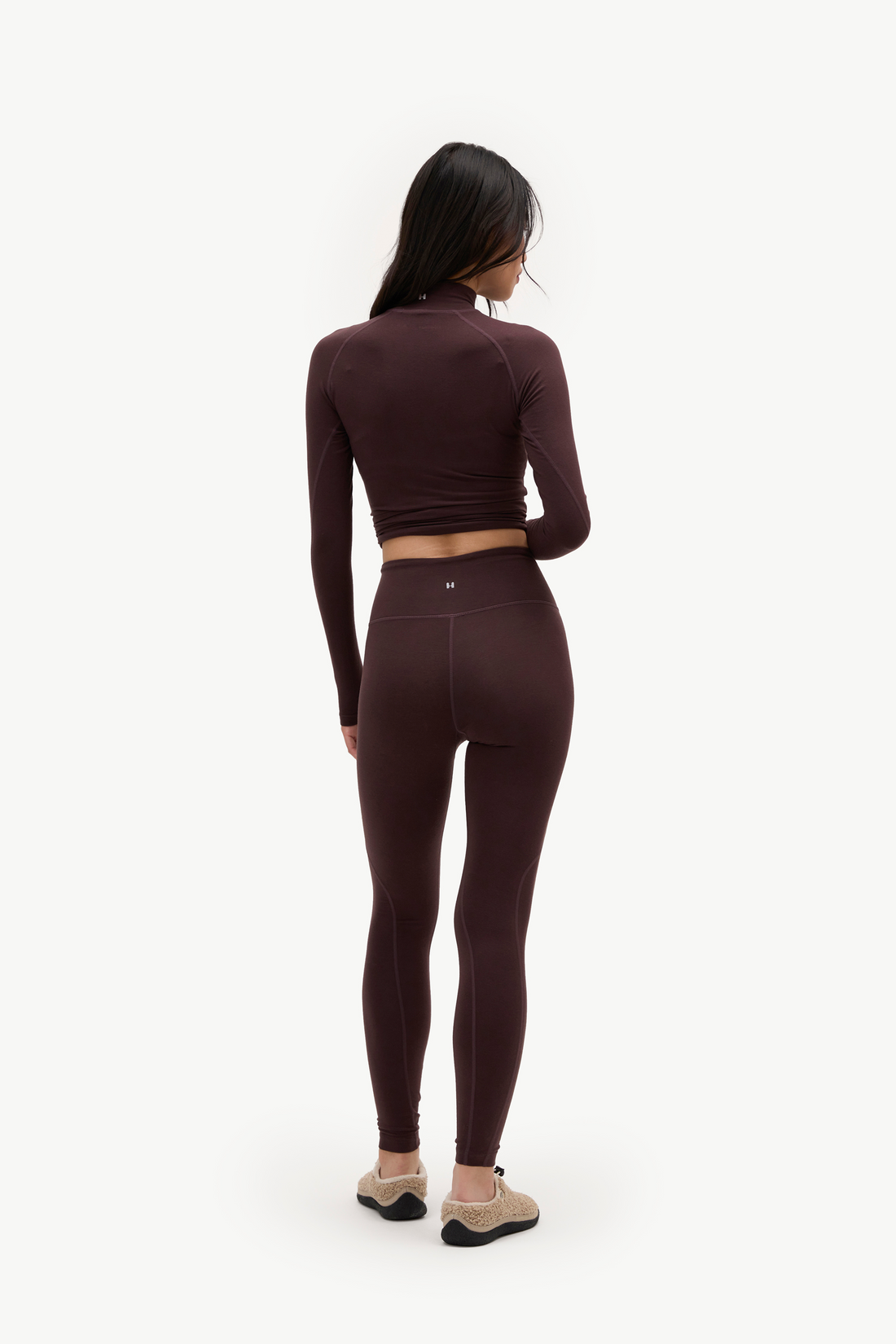 A woman is shown from behind wearing a matching dark brown athletic set consisting of a long-sleeve crop top and high-waisted leggings, paired with beige sneakers, against a plain white background.