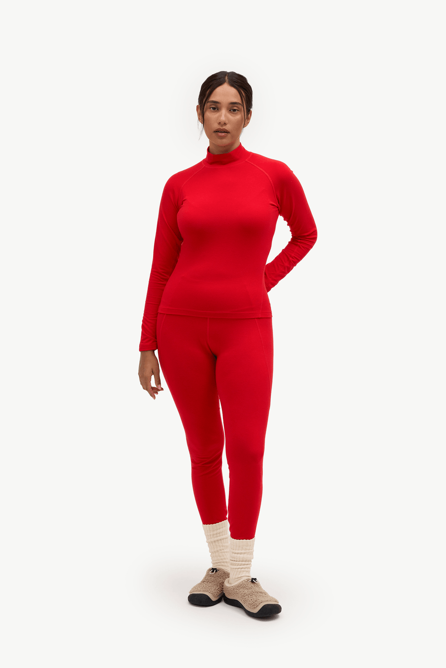A model wearing a matching bright red long-sleeve mock neck top and high-waisted leggings set, styled with cream ribbed socks and beige shoes, against a plain white studio background.