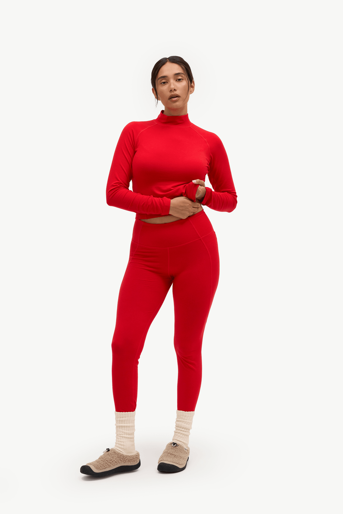 A woman modeling a matching red two-piece activewear set consisting of a long-sleeve mock neck crop top and high-waisted leggings, paired with beige socks and dark slip-on shoes, against a plain white studio background.