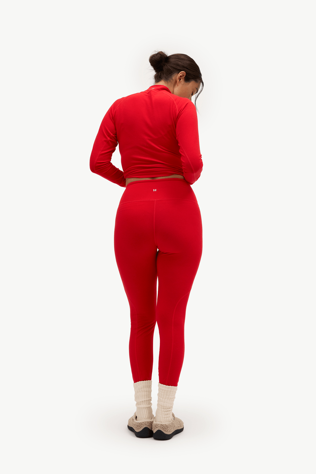 A woman is shown from behind wearing a matching red two-piece activewear set consisting of a long-sleeve crop top and high-waisted leggings, paired with beige socks and gray sneakers, against a plain white background.