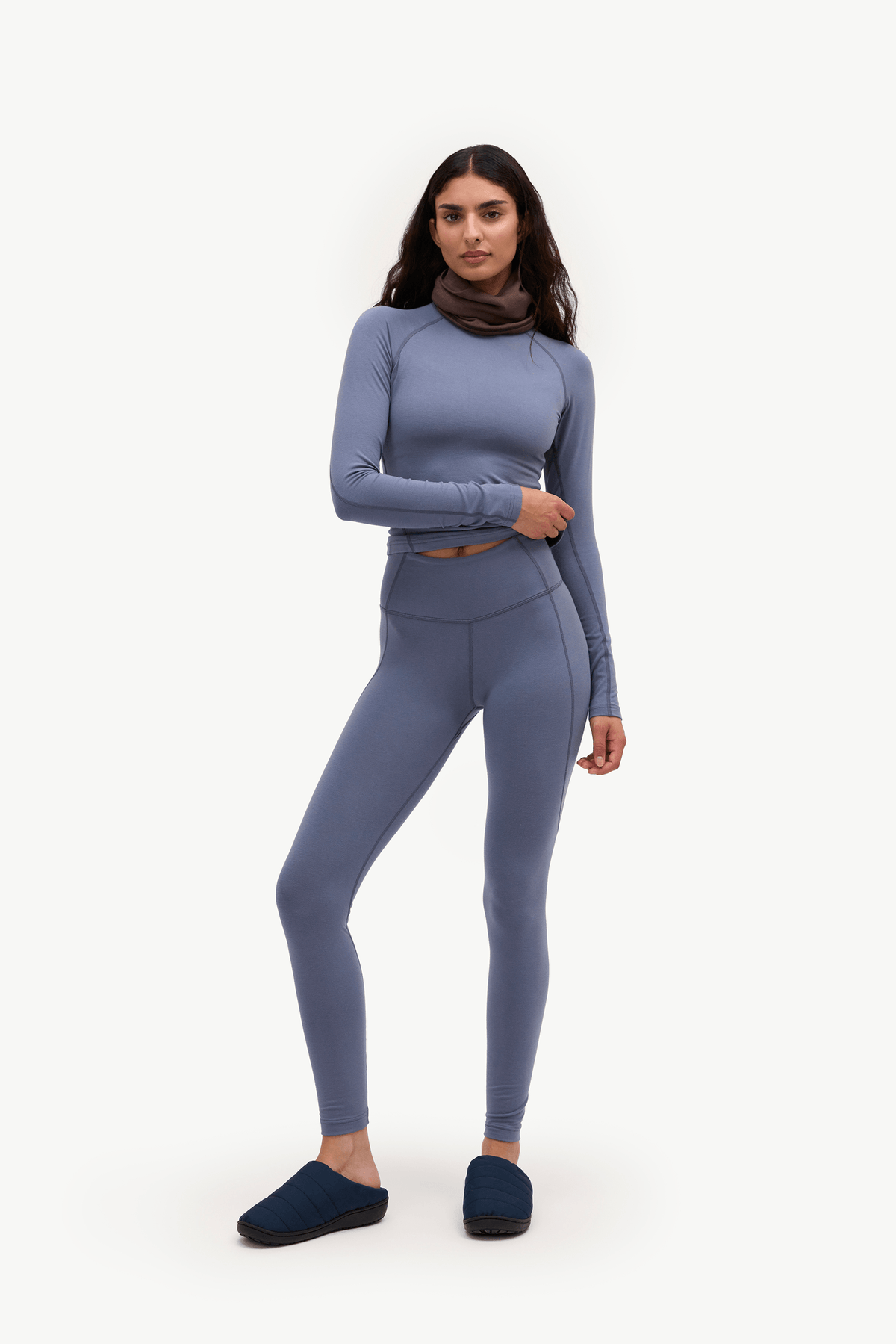 A woman modeling a matching two-piece activewear set in a dusty slate blue color, featuring a long-sleeve crop top and high-waisted leggings, paired with dark blue slippers against a plain white studio background.