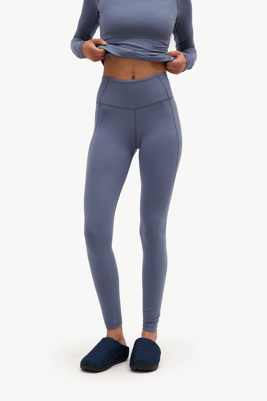 A model wearing high-waisted, full-length leggings in a muted slate blue-gray color, paired with a matching long-sleeve crop top and dark blue slip-on shoes, against a plain white studio background.