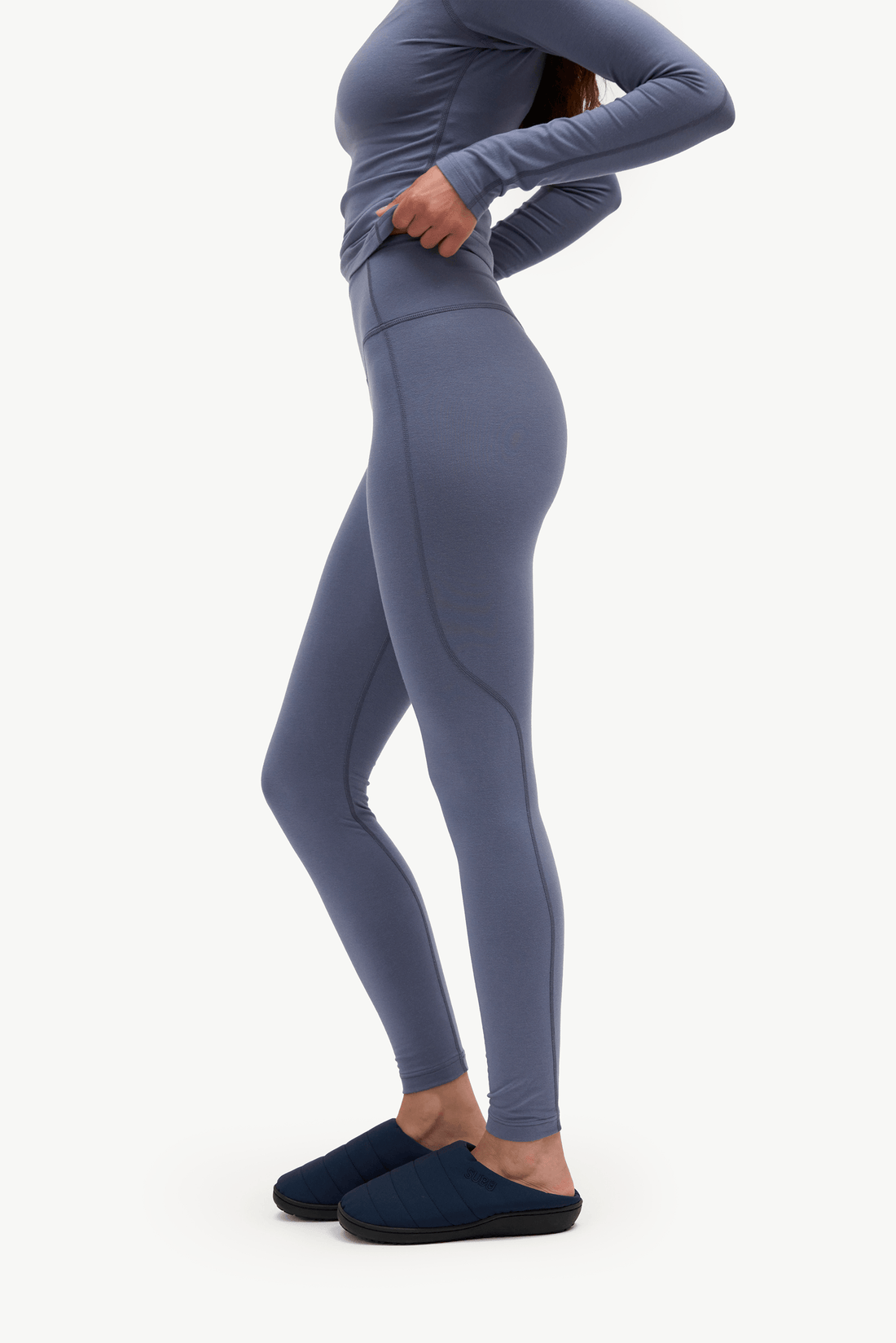 A model wearing a matching slate blue-gray long-sleeve top and high-waisted leggings, paired with dark navy slip-on slippers, shown from a side profile against a plain white studio background.