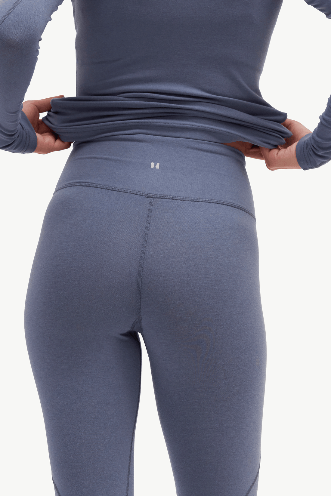 Close-up back view of a person wearing a matching slate blue-gray long-sleeve top and high-waisted leggings, pulling the top slightly away from the waist to show the fit and a small centered logo.