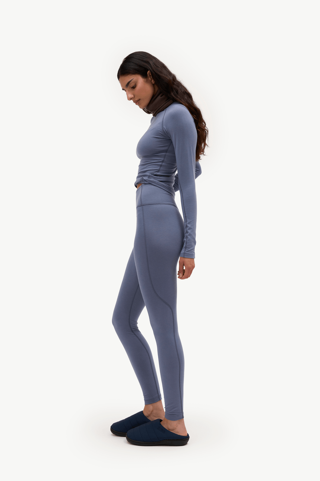 A woman in a side profile wearing a matching dusty blue-gray long-sleeve top and high-waisted leggings set, paired with dark navy slip-on slippers, against a plain white studio background.