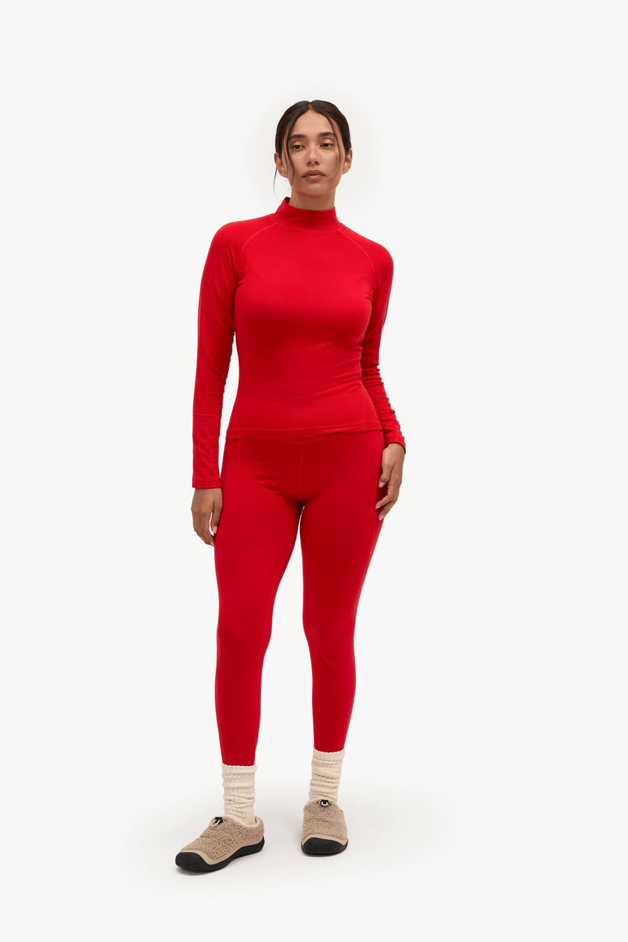 A model stands against a plain light background wearing a matching bright red long-sleeve mock-neck top and high-waisted leggings, paired with beige socks and dark slip-on shoes.