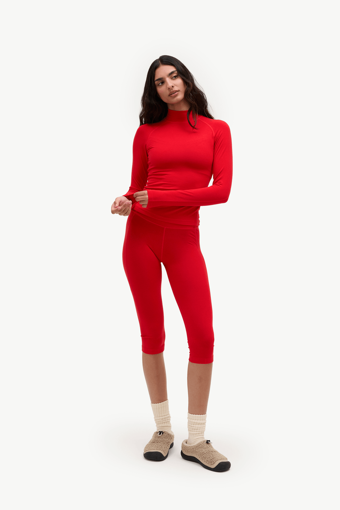 A woman modeling a matching red long-sleeve mock-neck top and capri-length leggings set, paired with cream socks and tan slip-on shoes, against a plain light gray studio background.