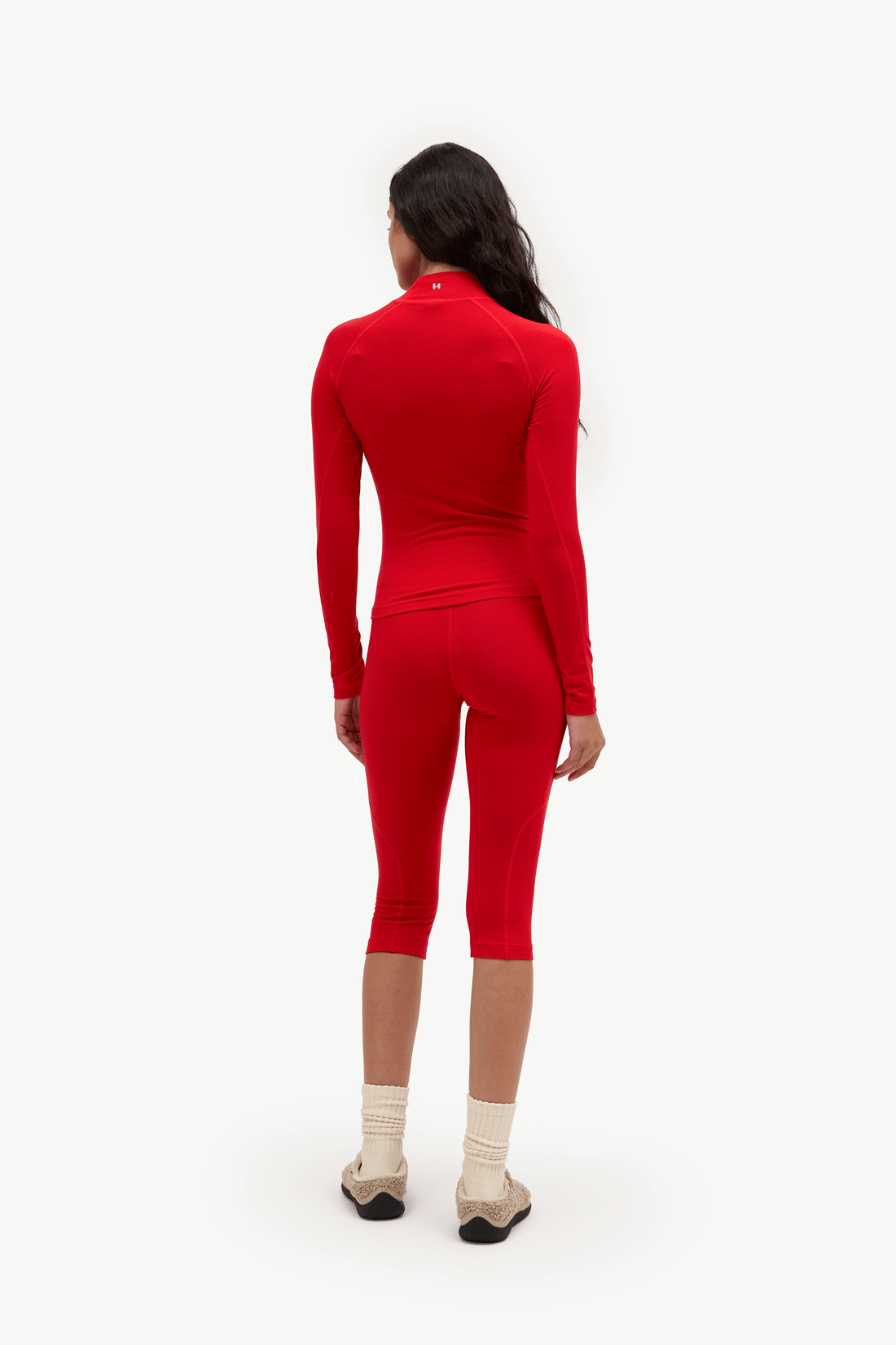 A woman viewed from the back wearing a bright red long-sleeve mock-neck top and matching red capri leggings, paired with cream socks and beige shoes, against a plain white studio background.