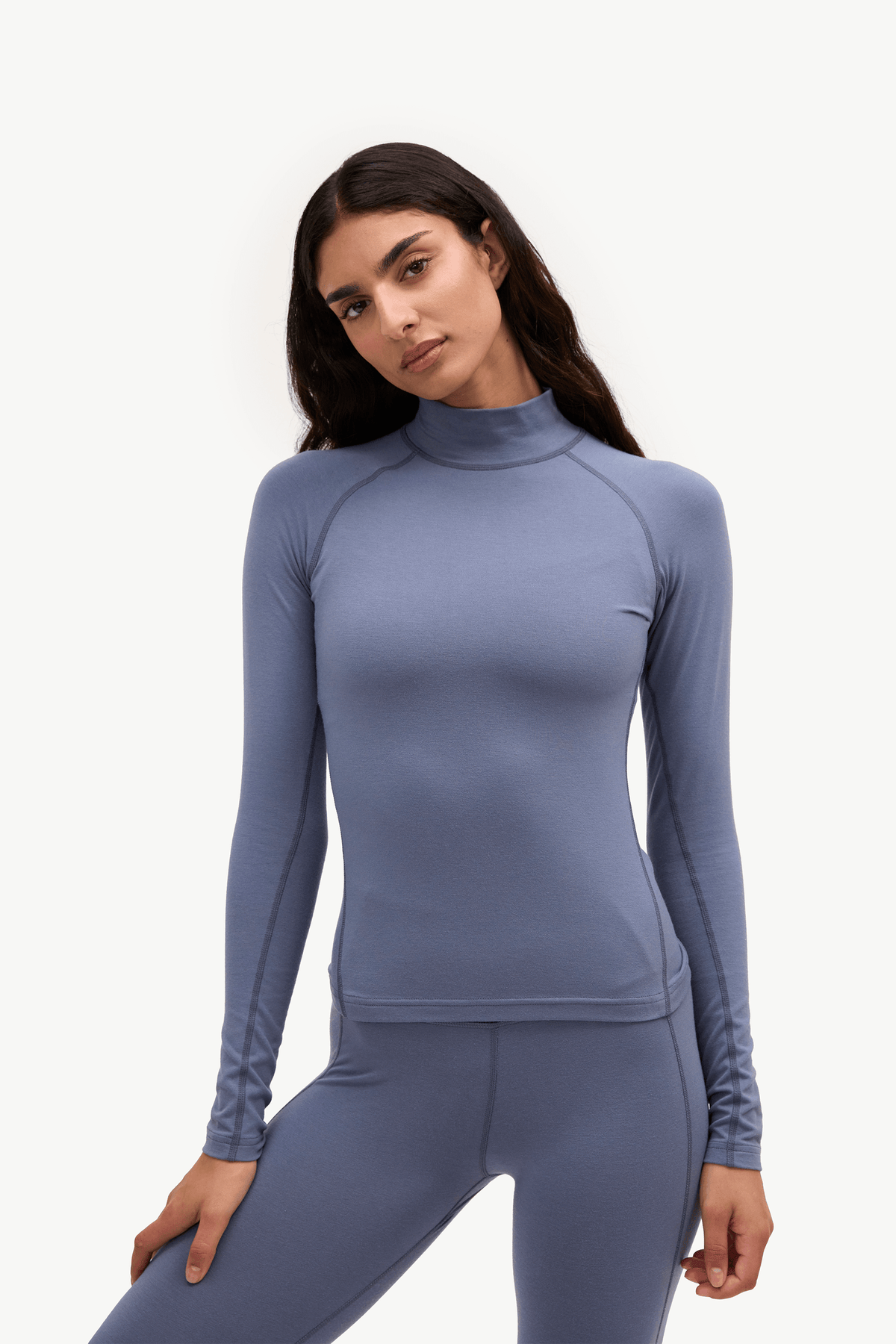 A woman modeling a matching long-sleeve mock neck athletic top and leggings set in a dusty periwinkle blue color, shot against a clean white studio background.