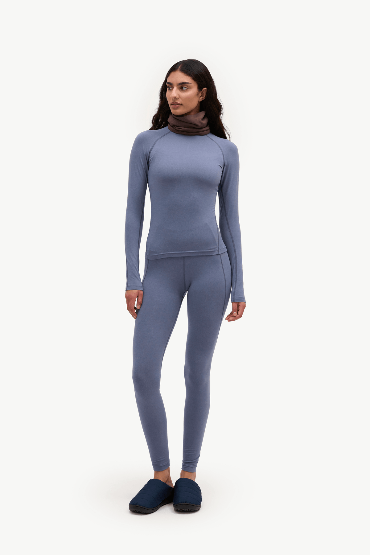 A woman modeling a matching two-piece athletic set consisting of a long-sleeve mock-neck top and high-waisted leggings in a dusty slate blue color, paired with dark navy slip-on shoes against a plain white background.