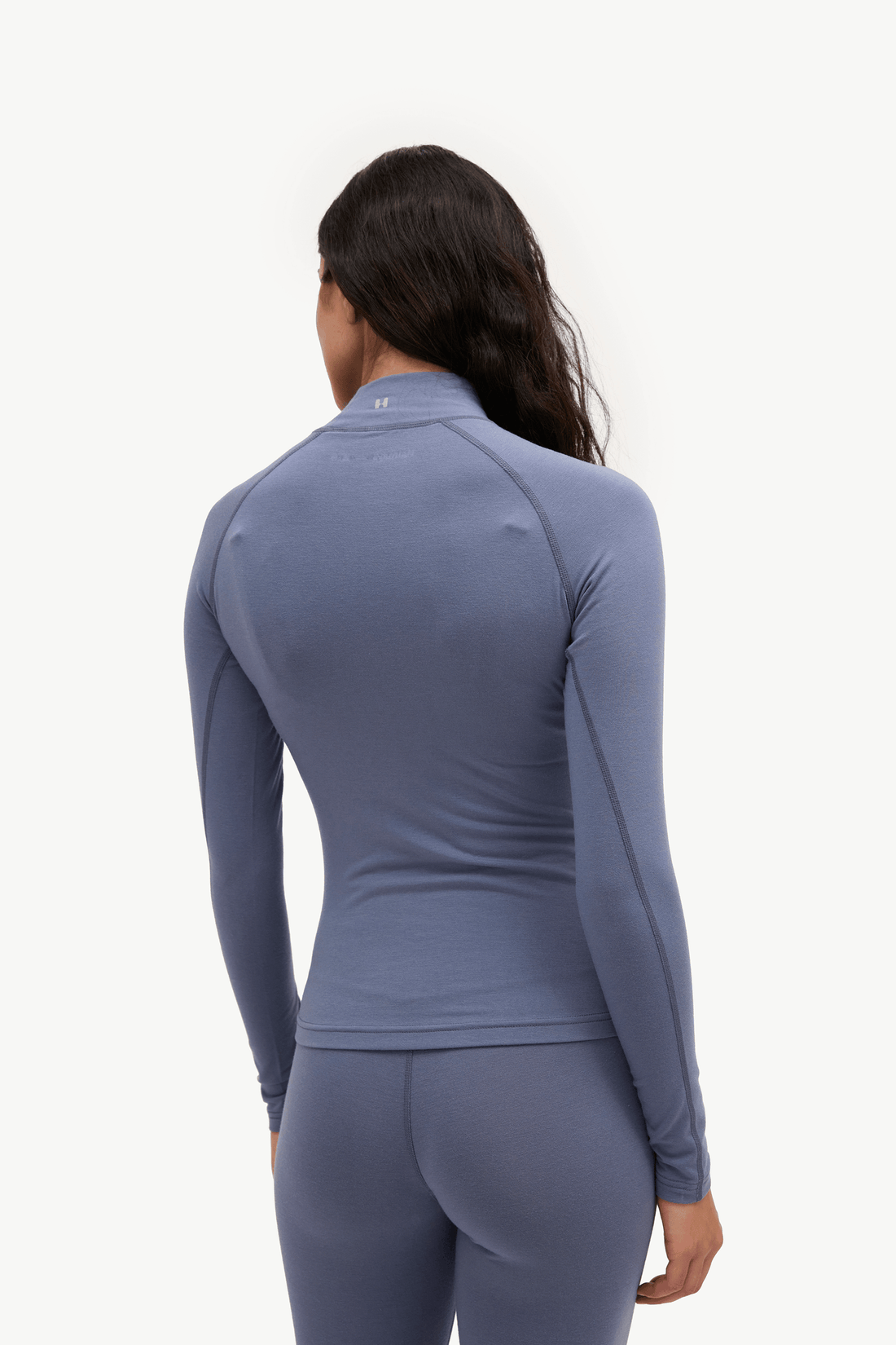 A woman shown from the back wearing a fitted long-sleeve athletic top and matching leggings in a muted dusty blue-gray color, against a clean white studio background.