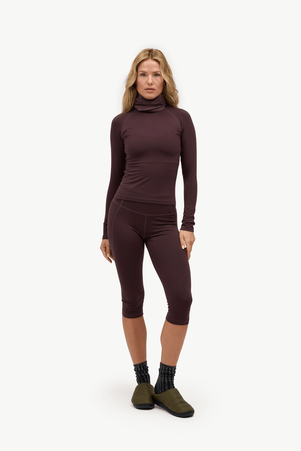 A woman modeling a matching dark plum-brown long-sleeve turtleneck top and capri leggings set, paired with dark socks and olive green shoes, against a plain white studio background.