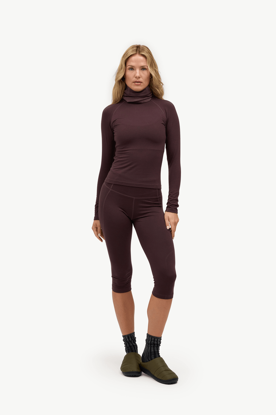 A woman modeling a matching dark plum-brown long-sleeve turtleneck top and capri leggings set, paired with dark socks and olive green shoes, against a plain white studio background.