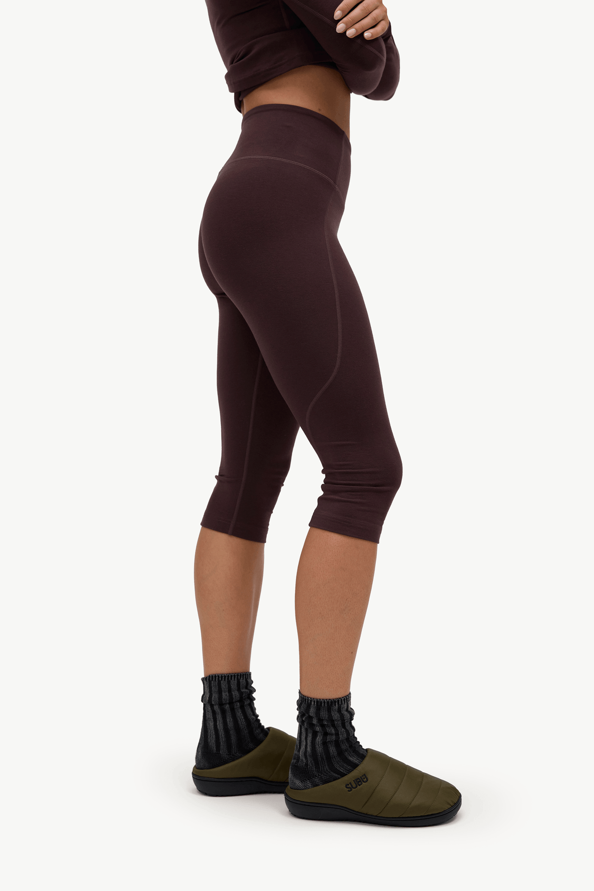 A model shown from the waist down wearing dark brown high-waisted capri leggings, black ribbed socks, and olive green slip-on shoes against a plain white background.