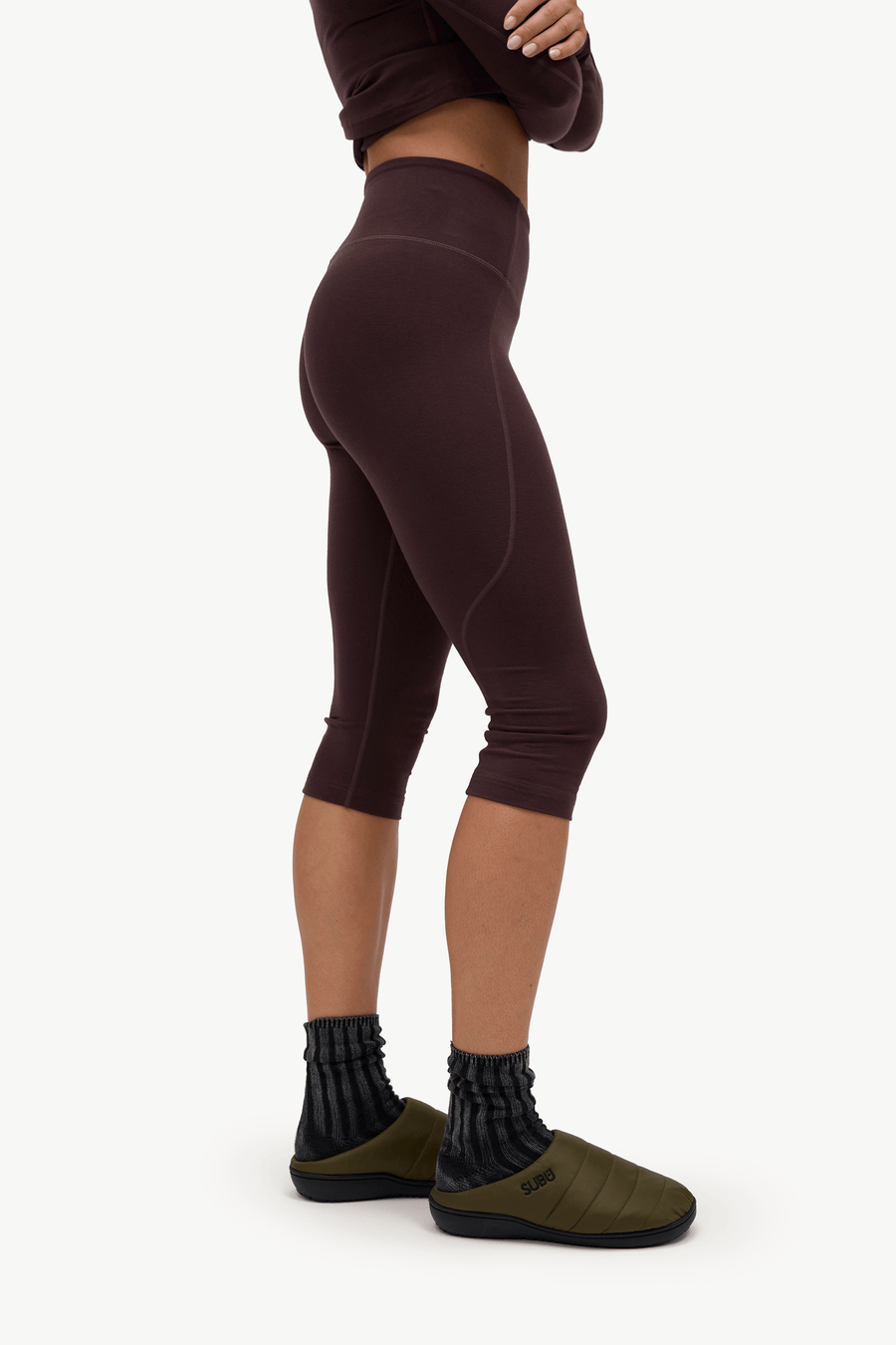 A model shown from the waist down wearing dark brown high-waisted capri leggings, black ribbed socks, and olive green slip-on shoes against a plain white background.