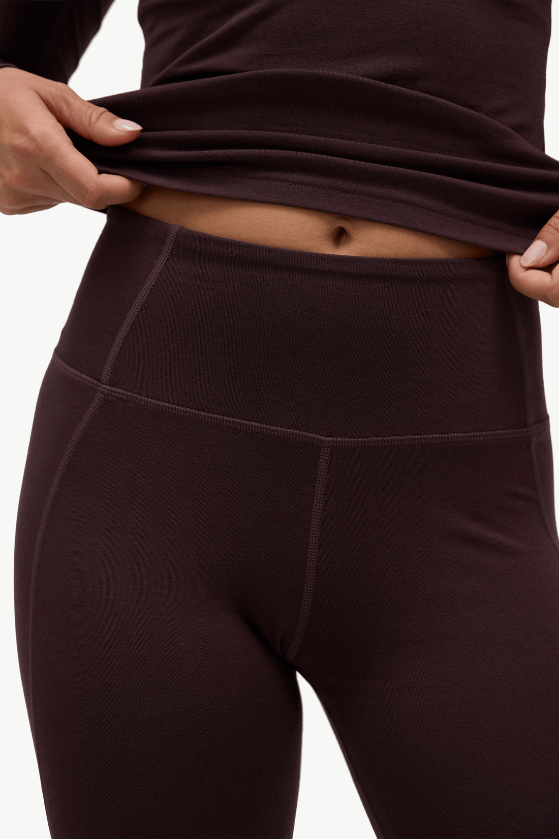 Close-up of a person wearing dark brown high-waisted leggings, lifting the hem of a matching top to show the waistband and fit against a plain light background.
