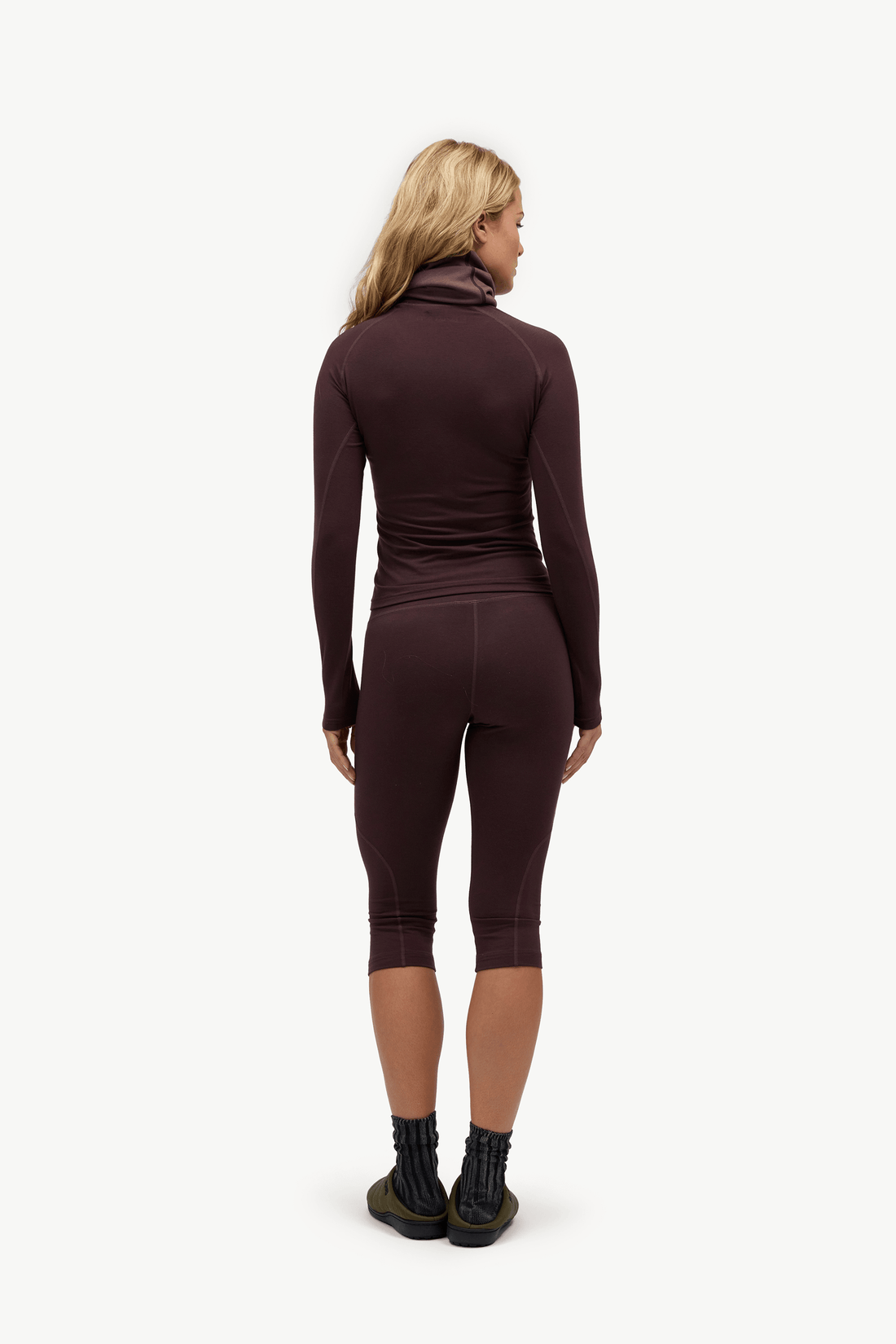 A woman is shown from the back wearing a matching dark plum-brown athletic set consisting of a long-sleeve mock-neck top and capri leggings, paired with black socks and running shoes, against a plain white background.