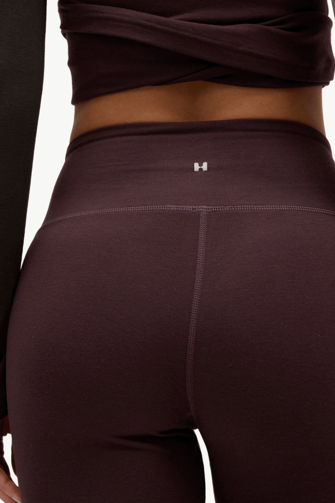 Close-up back view of a model wearing high-waisted leggings in a deep plum purple color, featuring a small silver 'H' logo on the center back waistband.