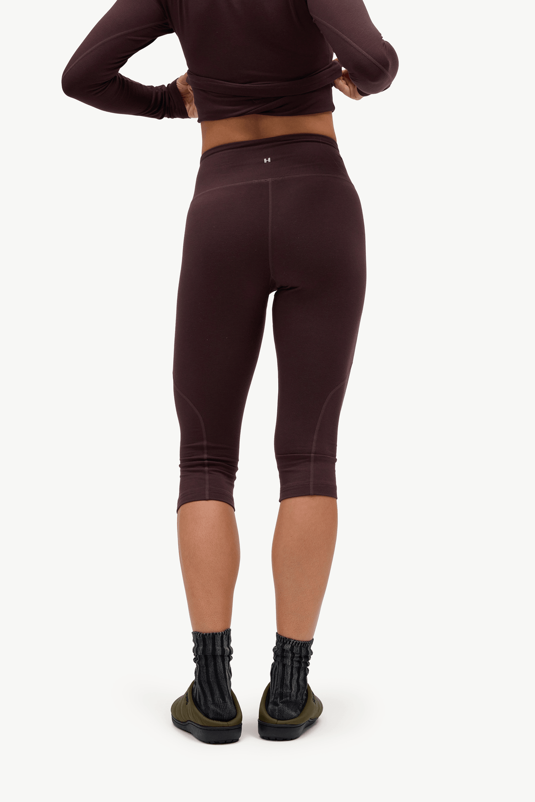 Rear view of a model wearing dark plum-brown high-waisted capri leggings, a matching long-sleeve crop top, and black socks against a clean white background.