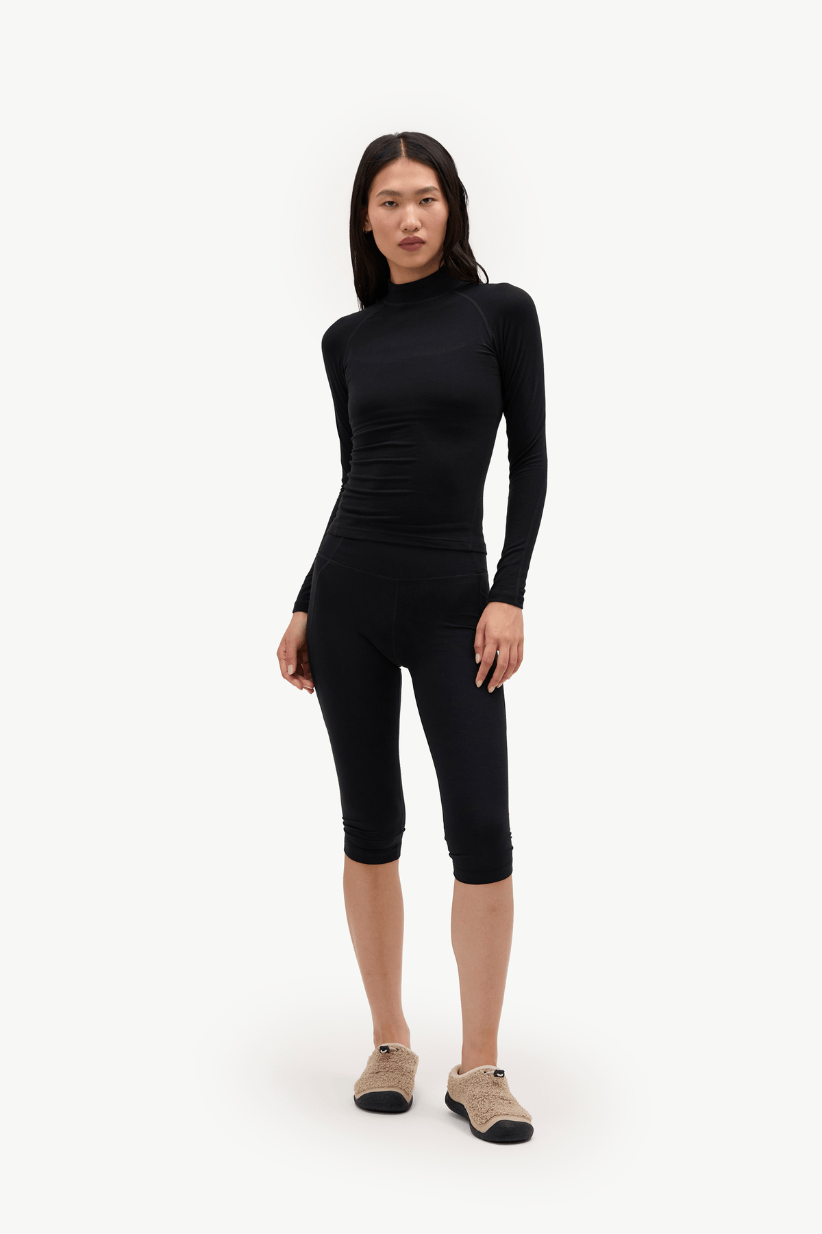 A model wearing a black long-sleeve mock neck top and matching black capri leggings, paired with beige slip-on shoes, against a plain white studio background.