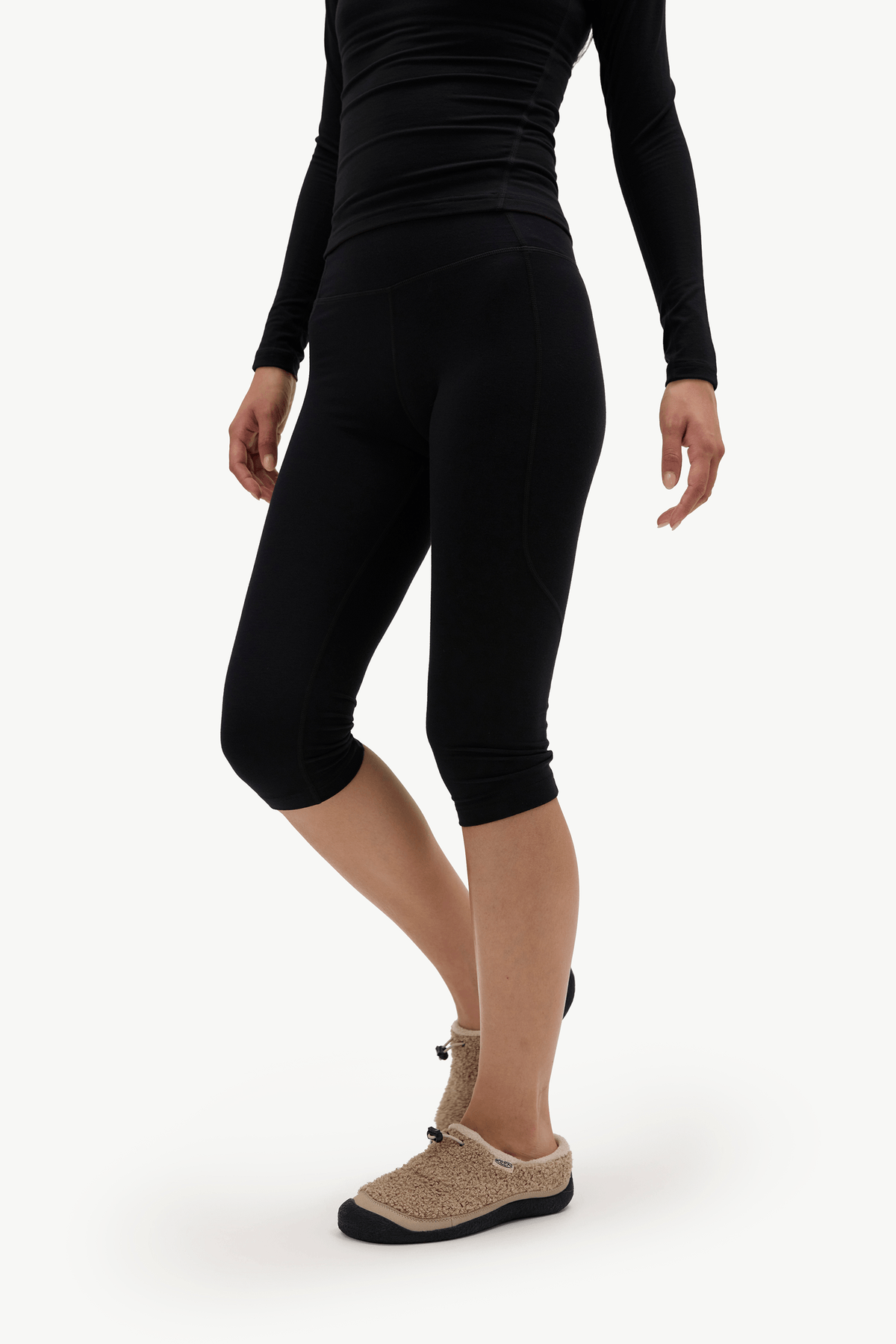 A model wearing black high-waisted capri leggings, a black long-sleeve top, and beige slip-on slippers against a plain white studio background.